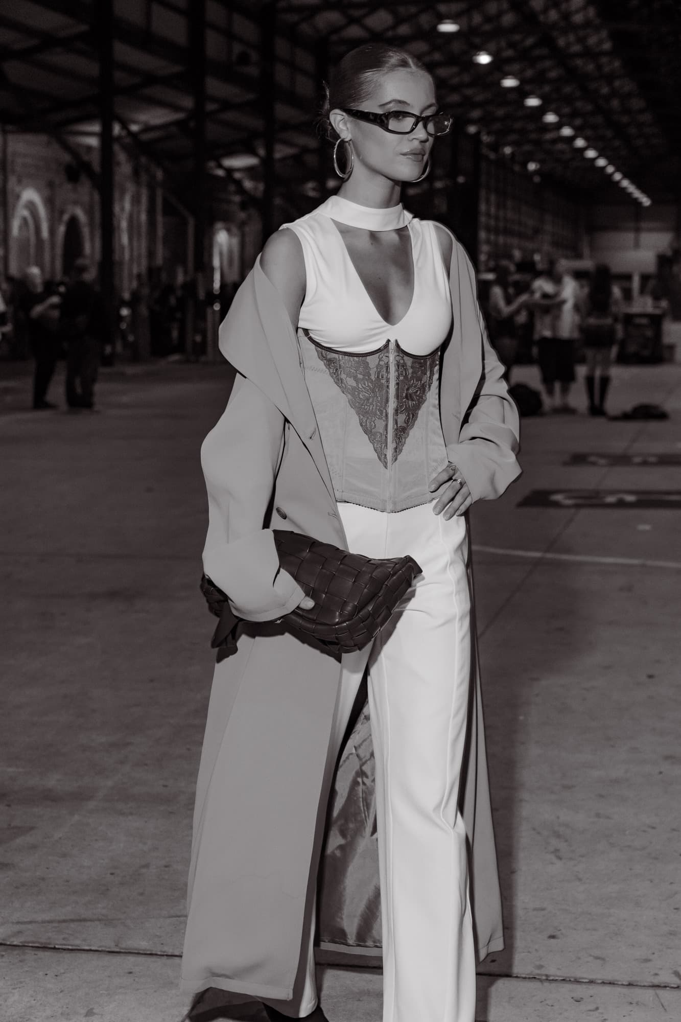 Black-and-white street-style portrait of a guest in a white lace corset, tailored coat and wide-leg trousers holding a woven leather clutch in a warehouse.