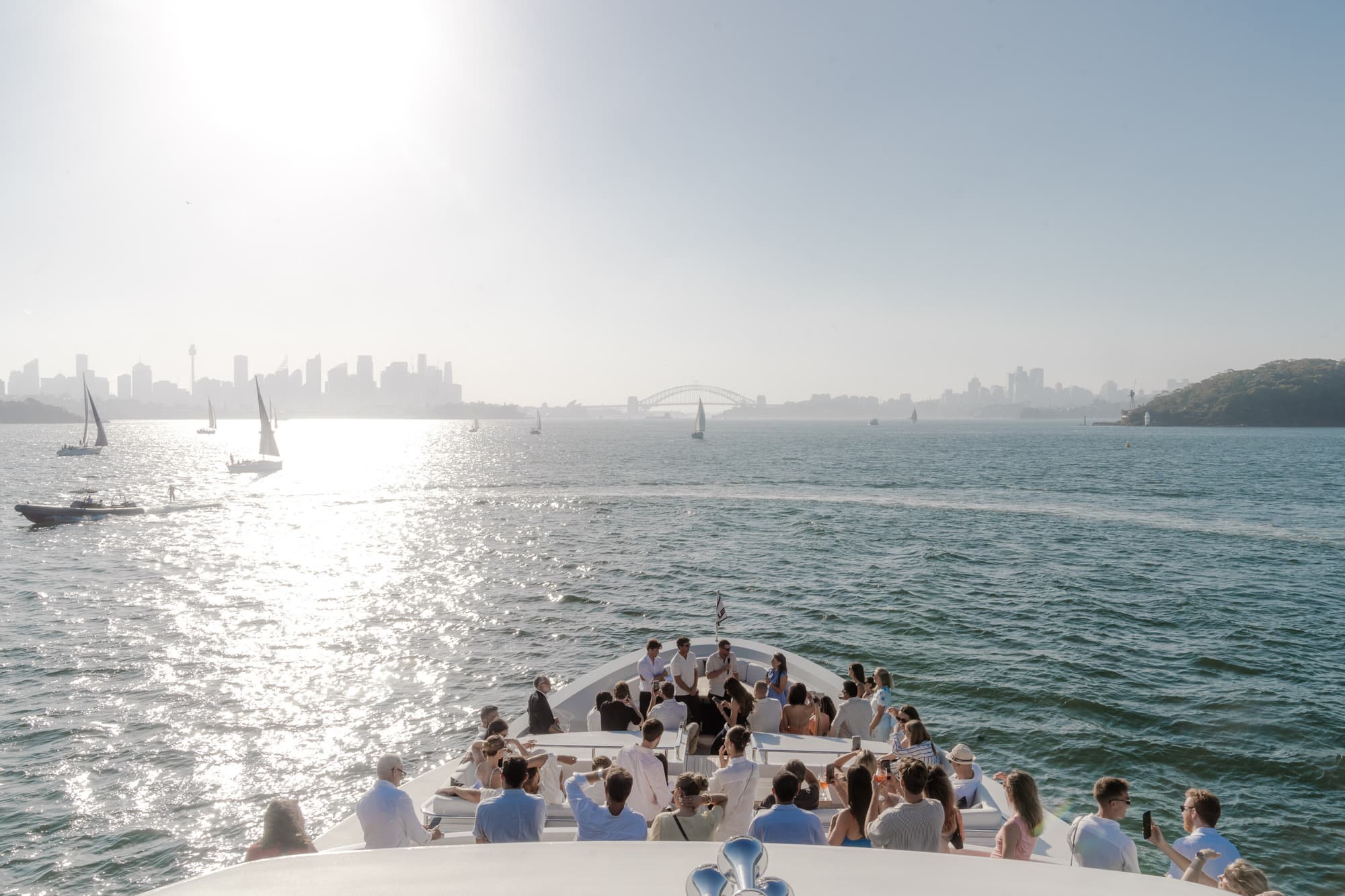 Guests in white gather on the bow of a boat on Sydney Harbour at golden hour, the Harbour Bridge and sailboats on the horizon.