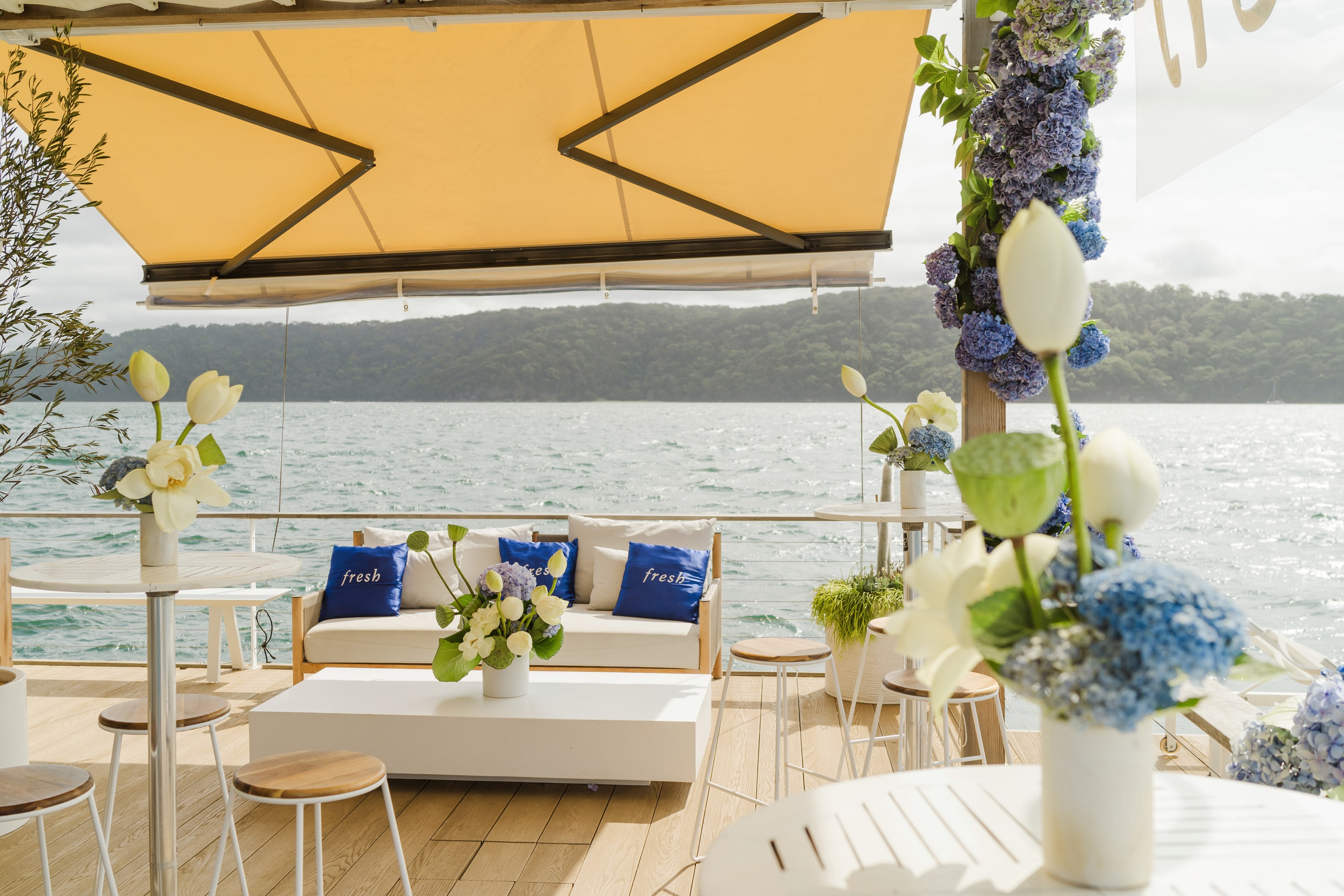 Fresh-branded blue cushions on a white lounge under a yellow awning, styled with white tulips and blue hydrangeas beside the water.