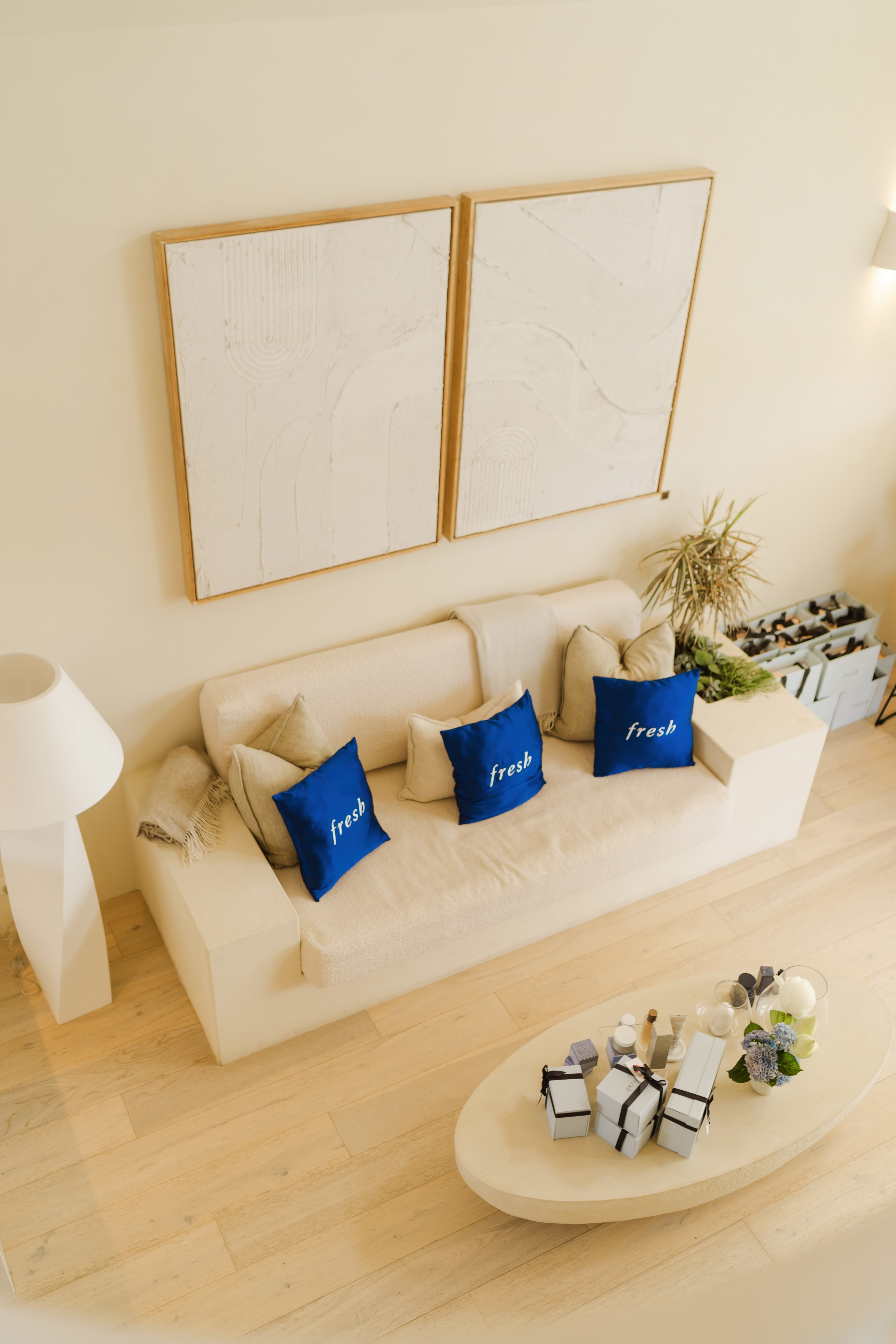 Cream lounge styled with blue Fresh cushions beneath abstract wall art, gift boxes and skincare on an oval coffee table.