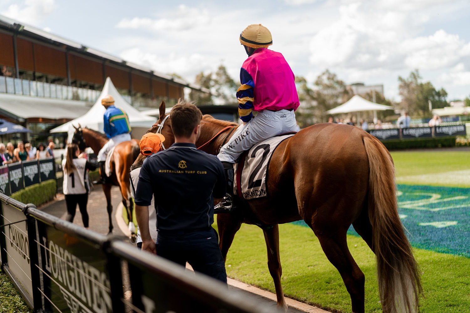 Jockeys on horseback are led along the mounting-yard rail at an Australian Turf Club raceday, Longines signage lining the track.