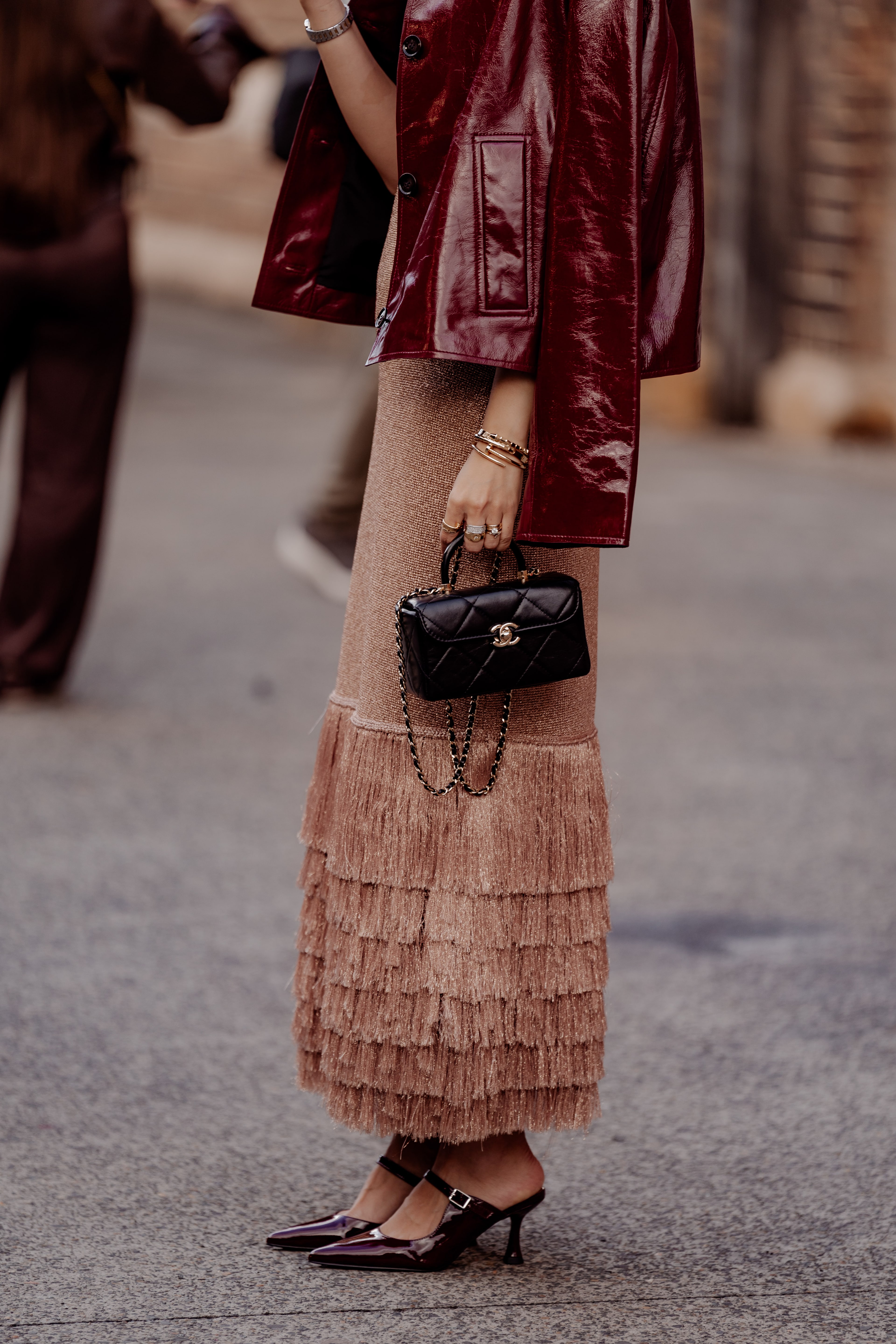 Street-style guest in an oxblood leather jacket and tiered fringed midi skirt holds a black quilted Chanel mini bag outside a fashion show.