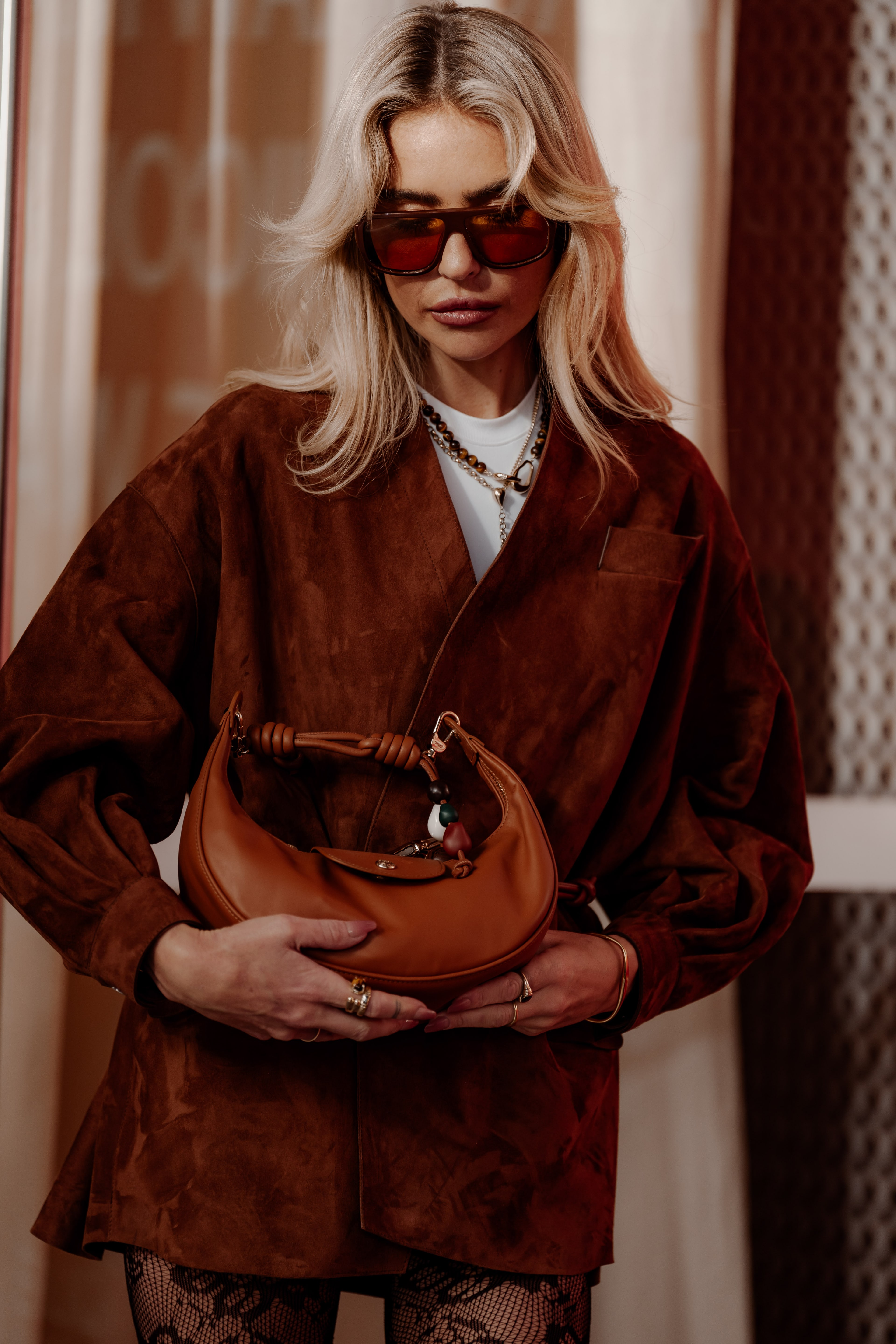 Blonde guest in tinted sunglasses and an oversized brown suede blazer cradles a tan leather shoulder bag at Australian Fashion Week.