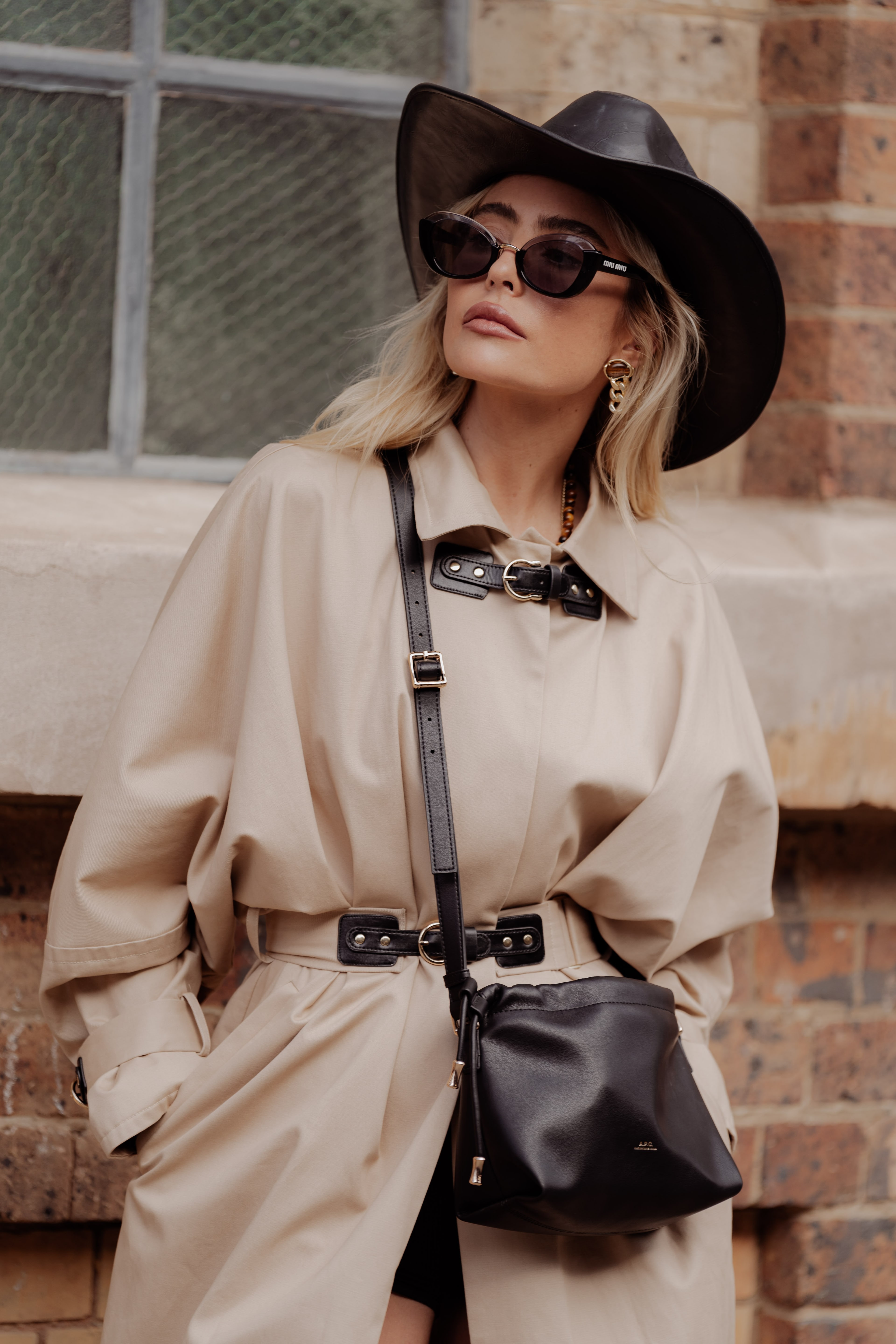 Blonde guest in a black wide-brim hat, sunglasses and belted beige trench coat with a black leather crossbody bag against a brick wall.