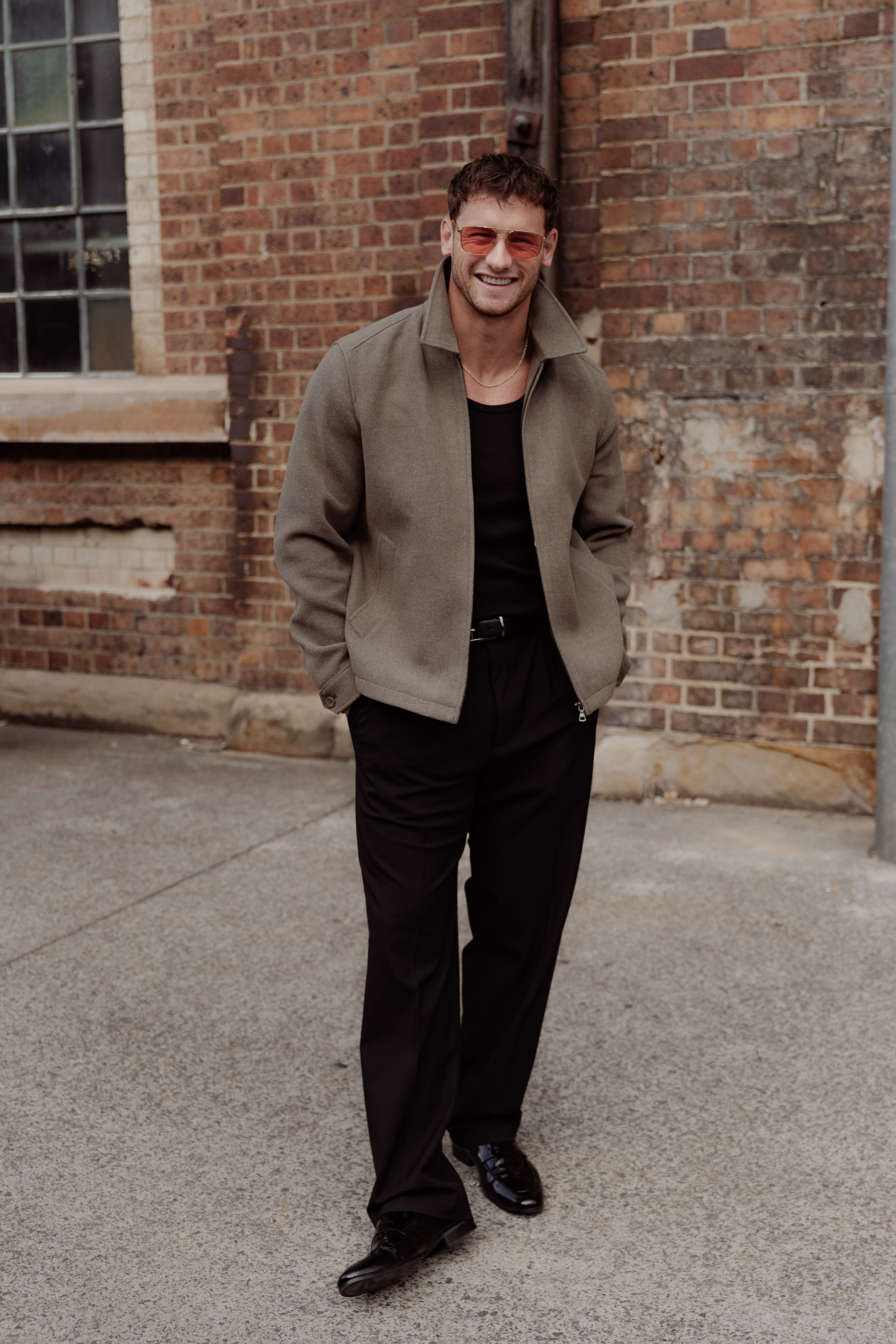 Smiling man in tinted sunglasses, an olive zip jacket, black tee and black trousers stands hands-in-pockets before a weathered brick building.