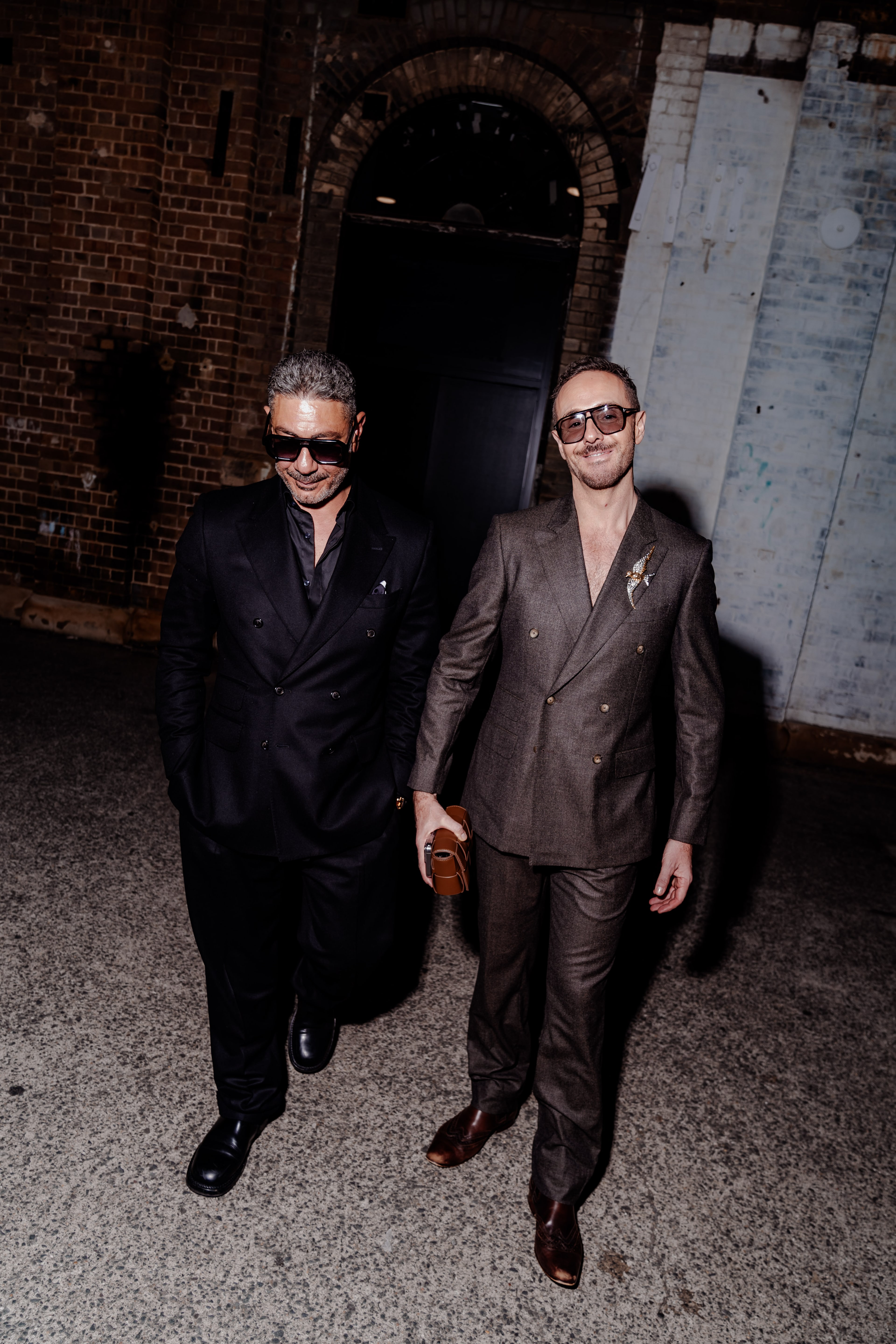 Two men in tailored black and brown suits with sunglasses walk together through a dim brick industrial venue at night.