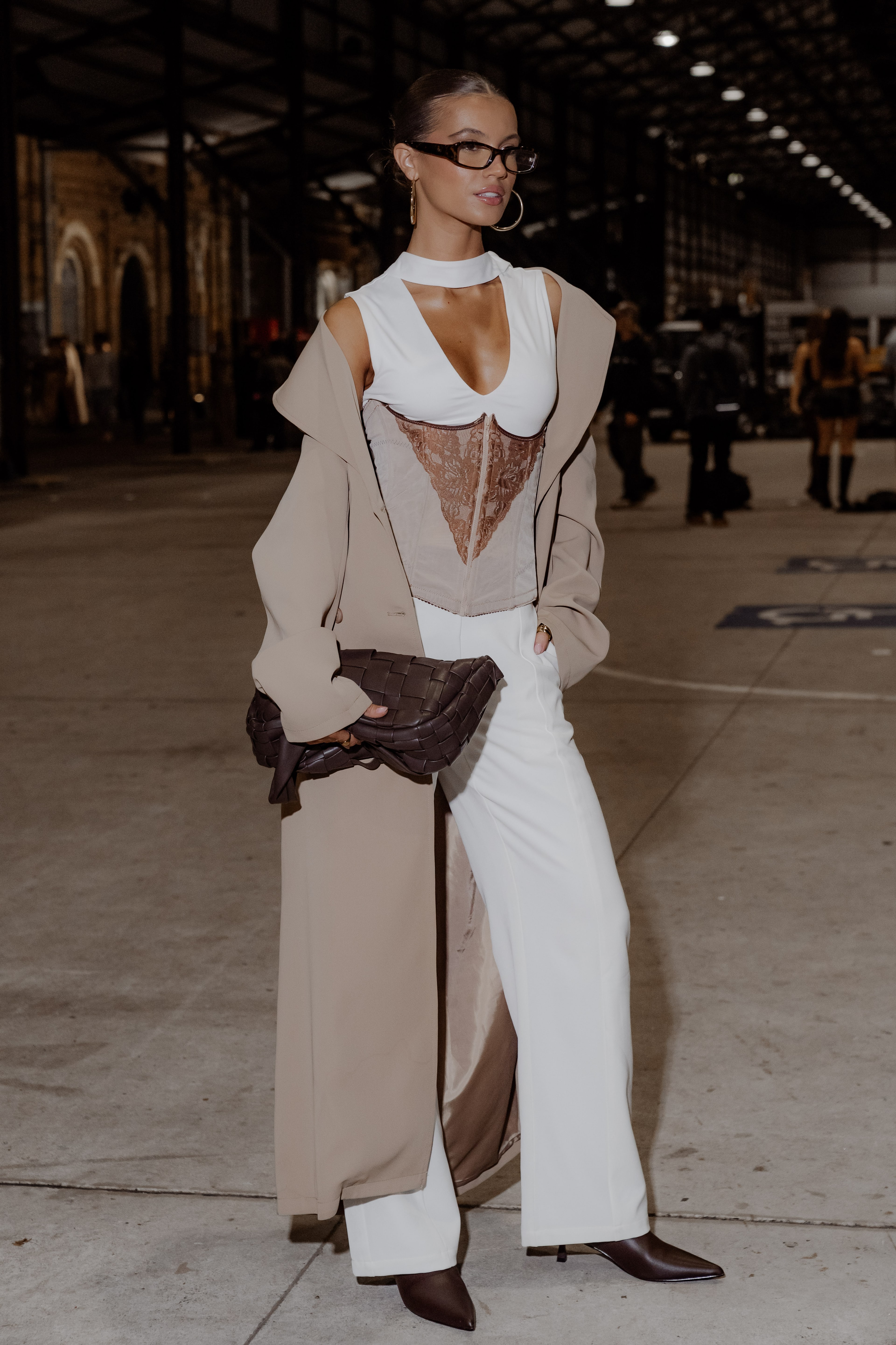 Guest in glasses wears a beige duster coat, sheer white corset top and wide white trousers, holding a brown clutch inside a show venue.