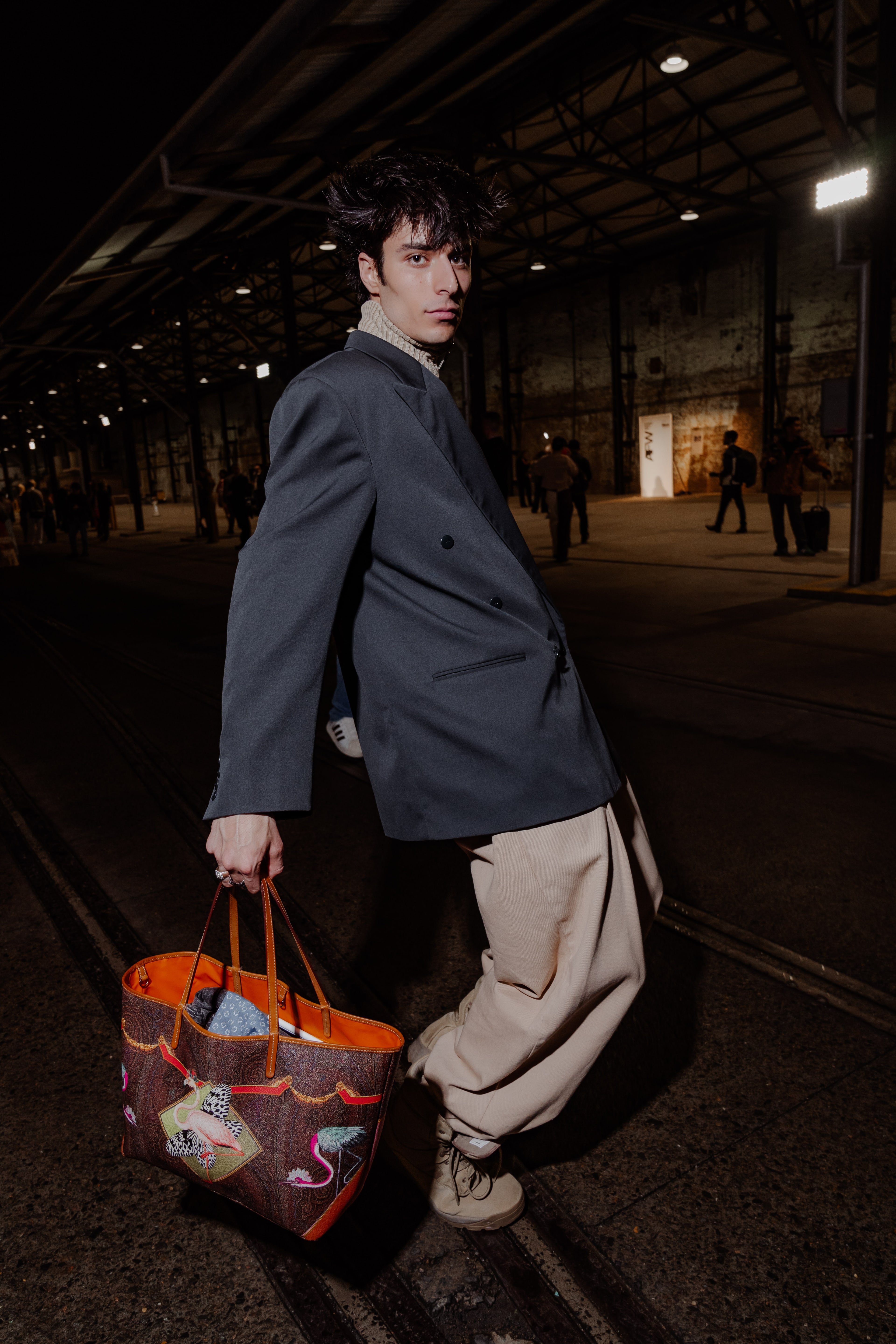Young man with tousled hair in an oversized navy blazer and beige trousers carries a printed orange tote across a dim industrial hall.