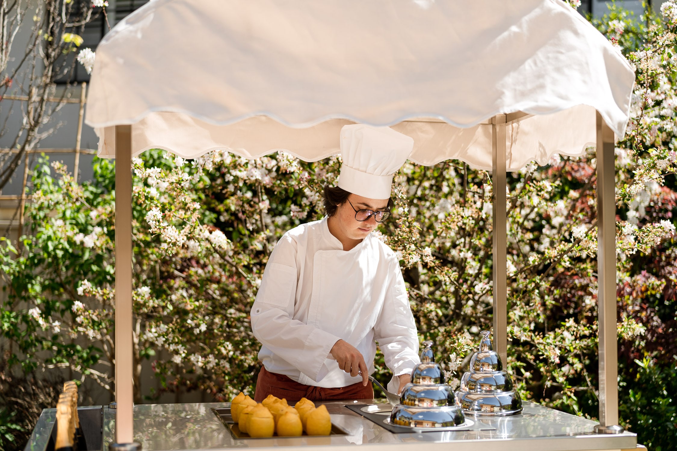 A chef in white uniform and toque slices at a canopied Pepi's cart with hollowed lemons and silver domes, backed by blossoming spring branches.