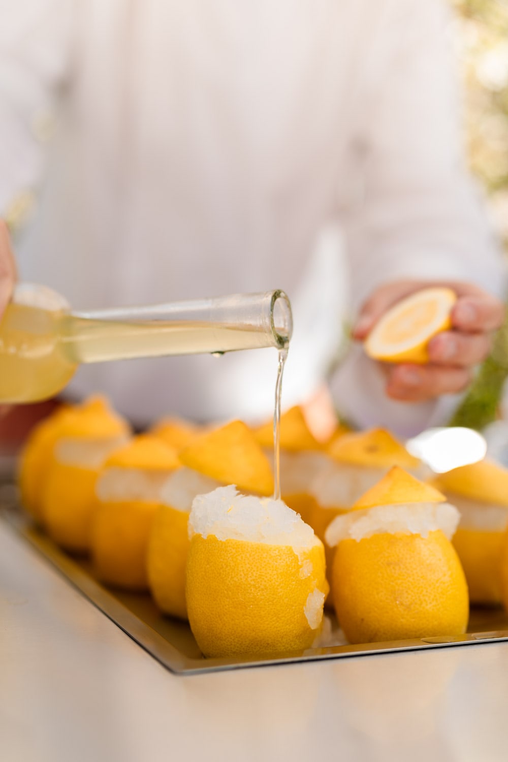 Close-up of pale liquor poured from a bottle into a tray of hollowed lemon shells filled with lemon sorbet, a halved lemon held behind.