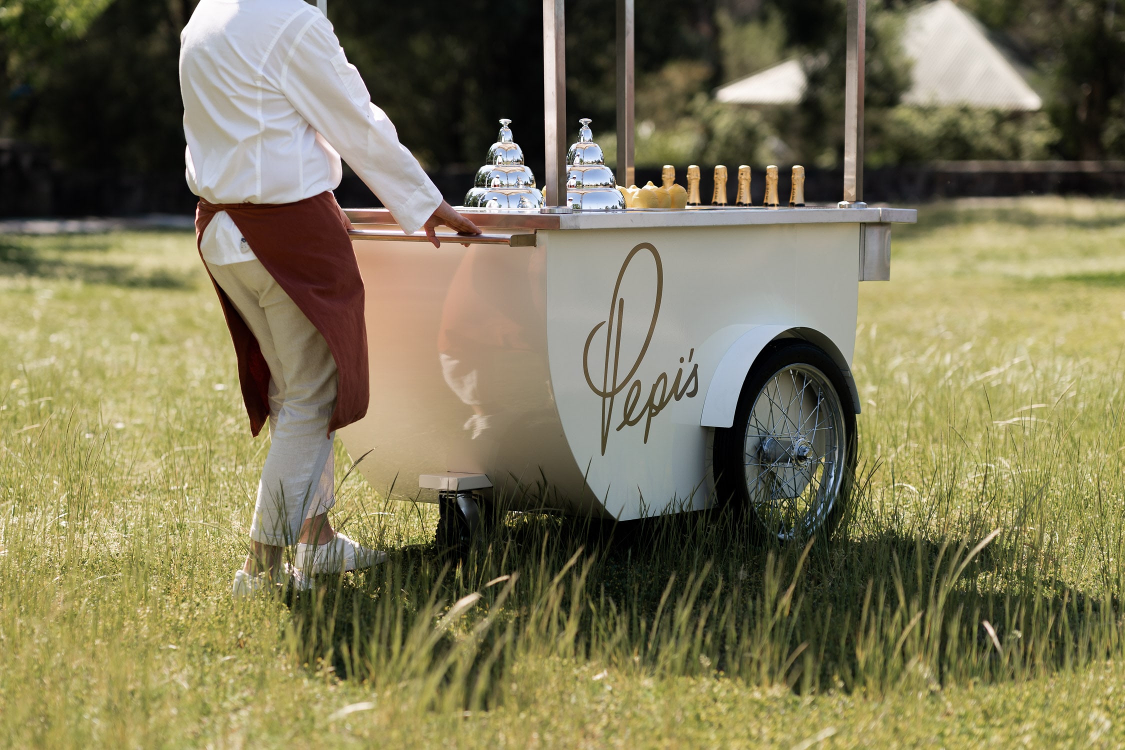 A server in white shirt and rust apron pushes the branded Pepi's cart, topped with silver domes and prosecco bottles, across a sunlit grassy field.
