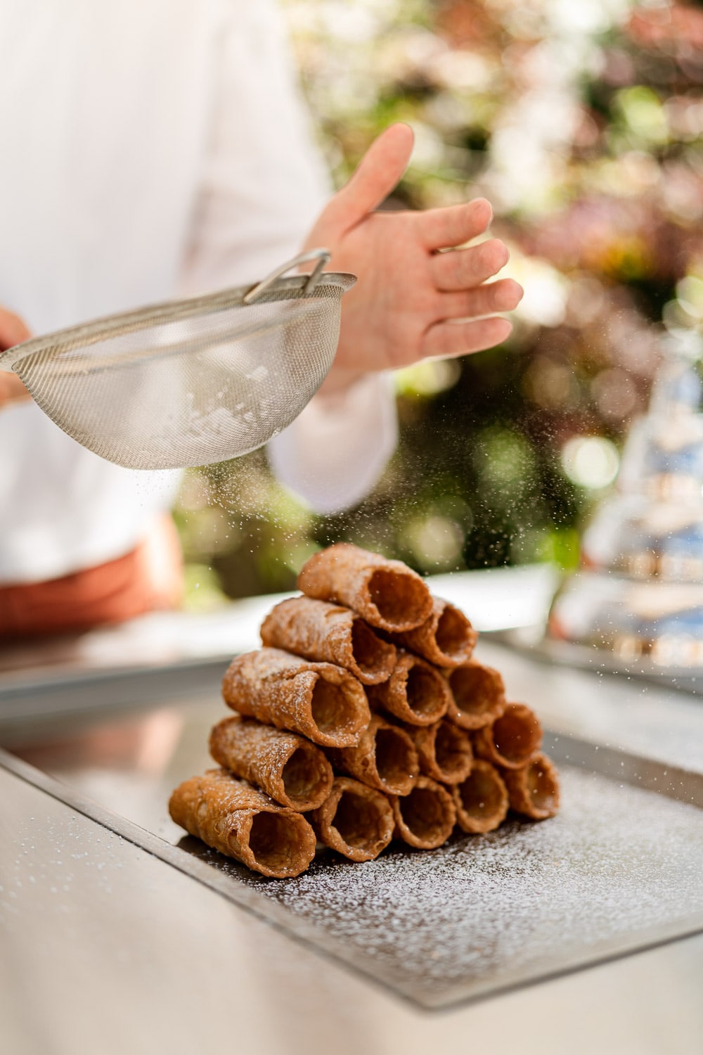 A chef dusts icing sugar through a sieve over a neatly stacked pyramid of cannoli shells on a metal tray outdoors.