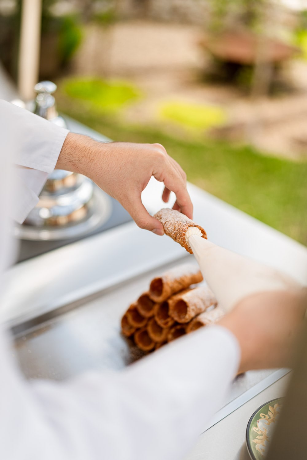 Close-up of a chef's hands piping cream filling into a sugar-dusted cannoli beside a row of stacked shells on a steel cart.