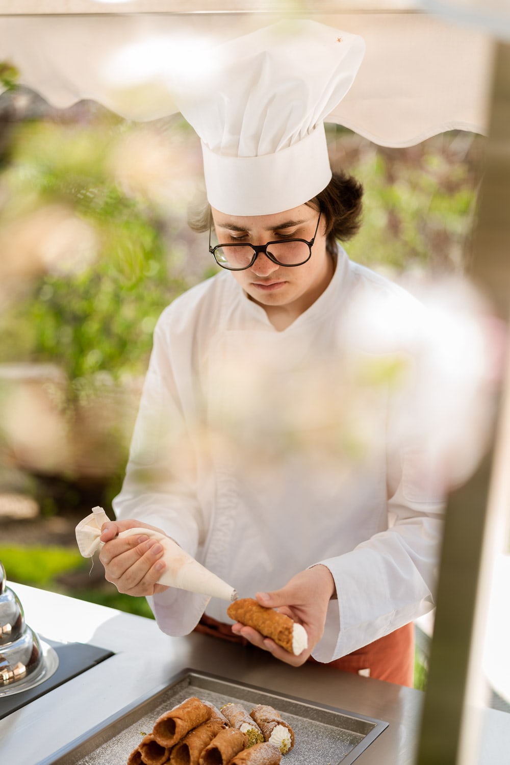 A young chef in white toque and glasses concentrates while piping cream into a cannoli shell over a tray of finished pastries.