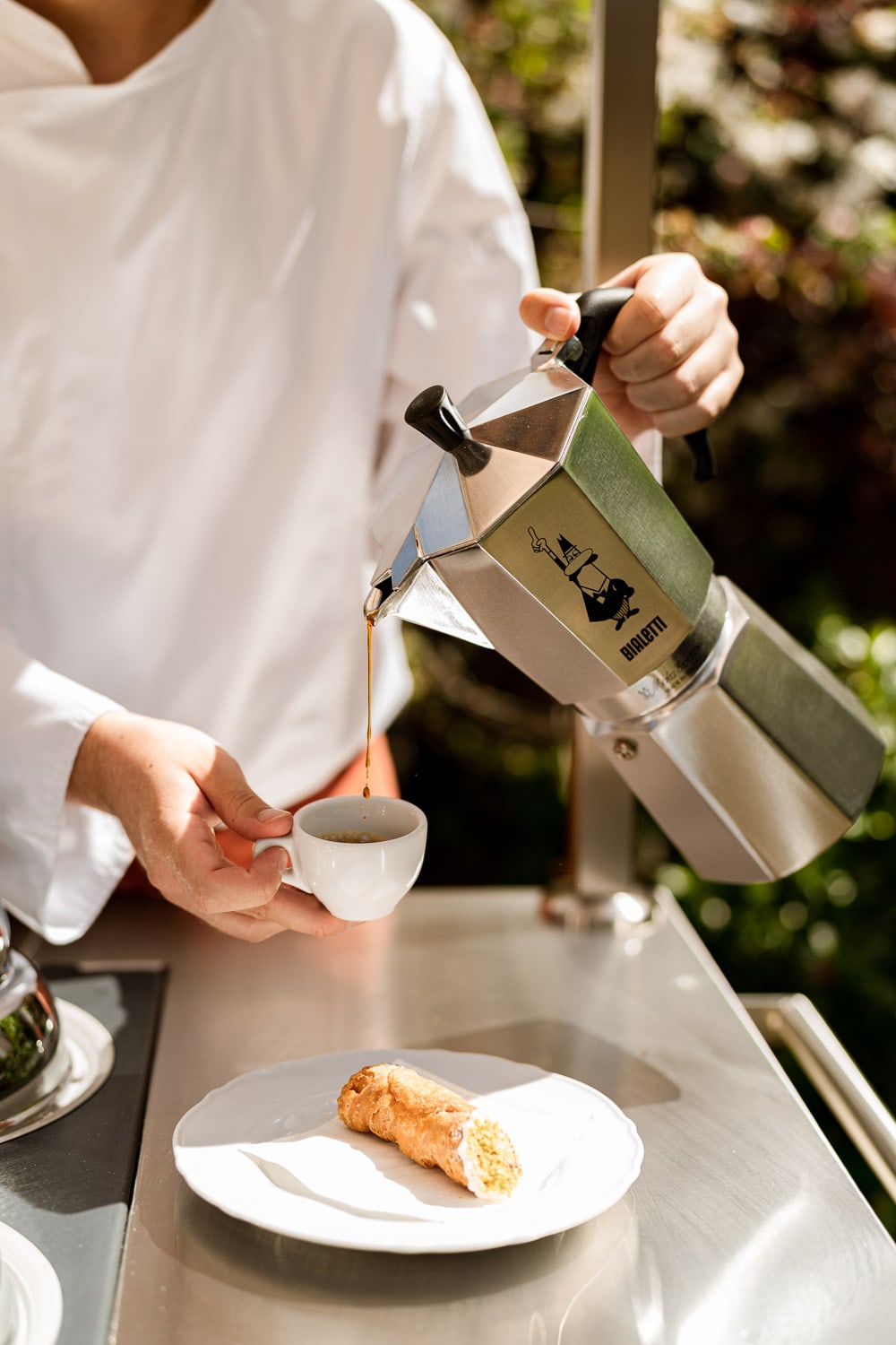 A server pours espresso from a Bialetti stovetop pot into a small white cup, with a filled cannoli plated on the steel cart.