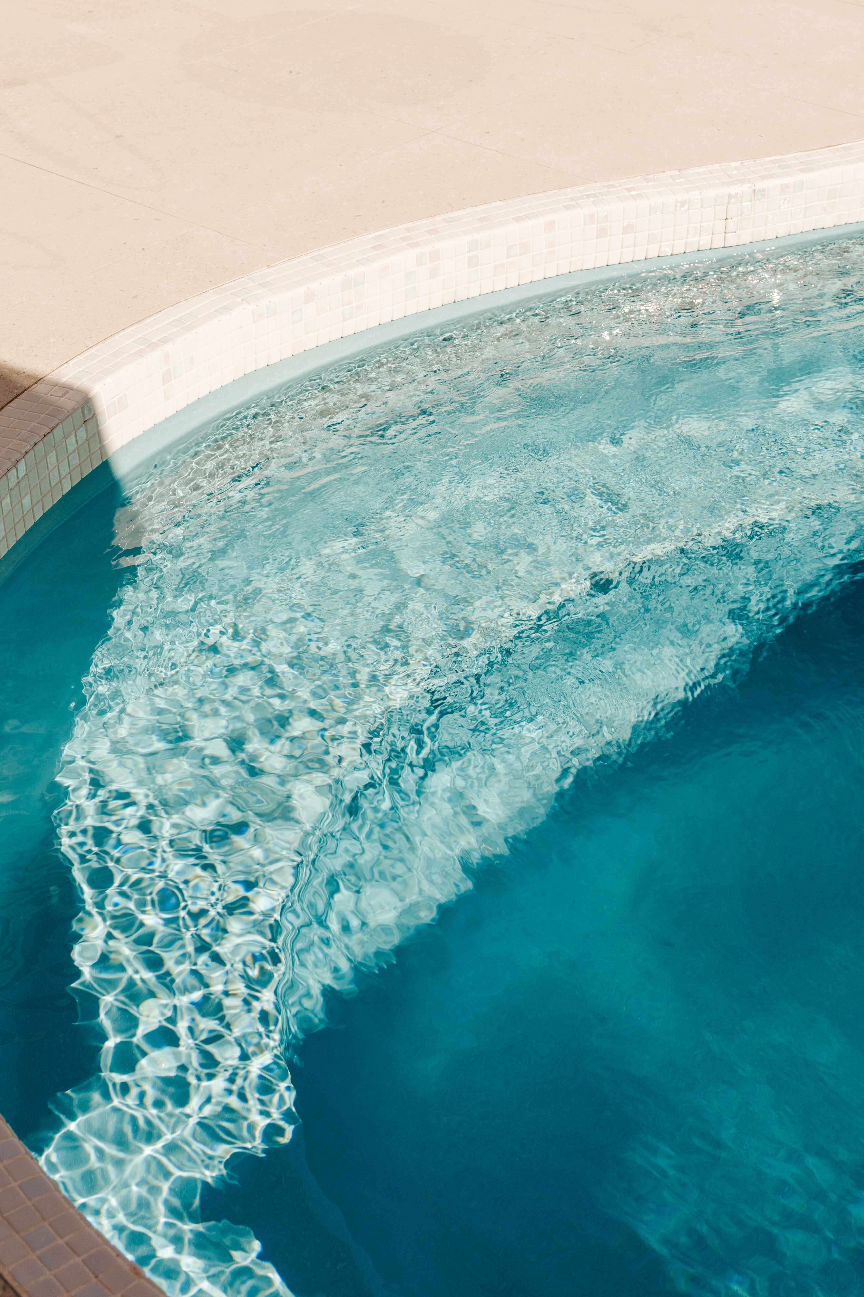 Sunlit turquoise pool water ripples along a curved tiled edge beside a smooth sand-toned deck.