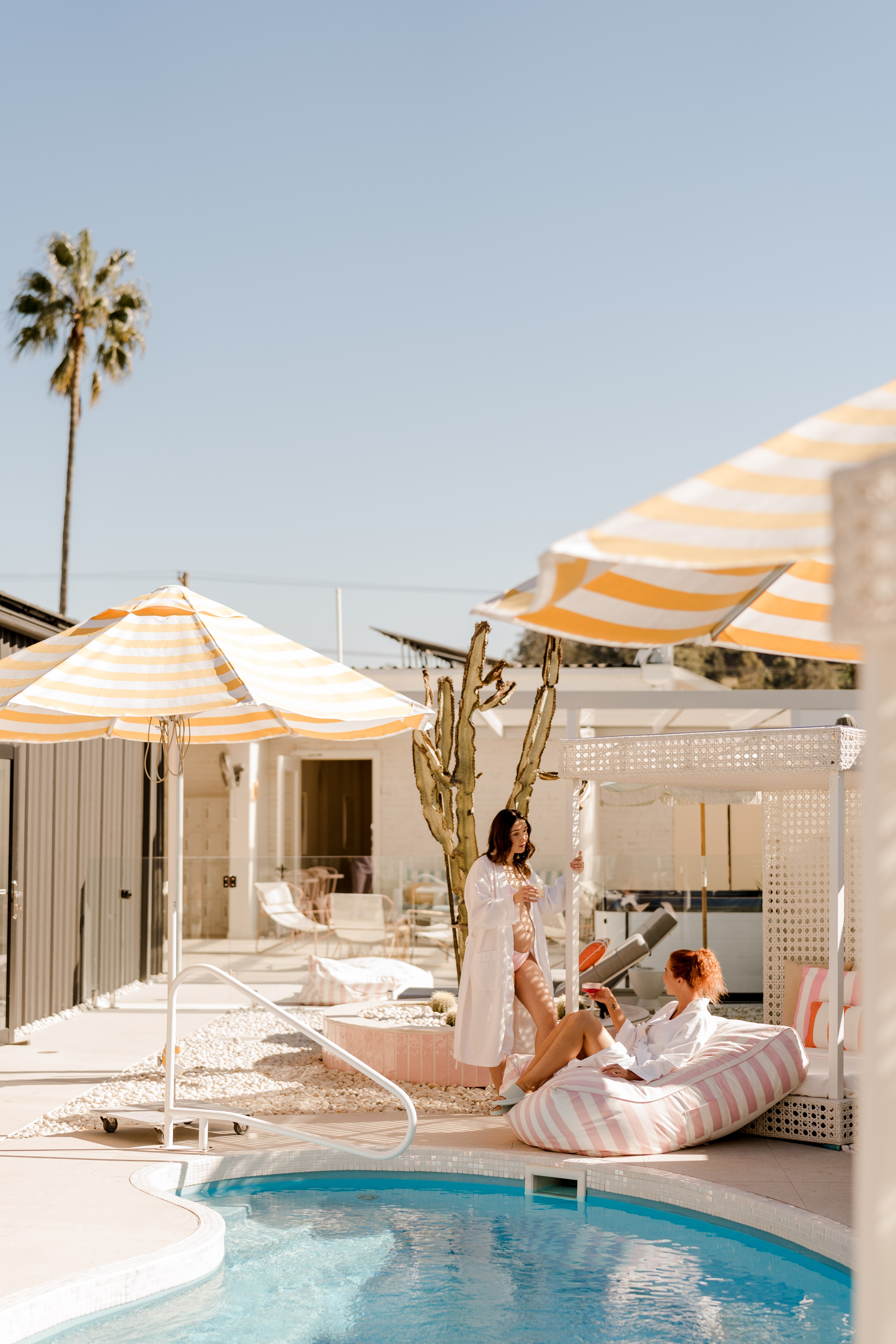 Two women in white robes relax by the pool under striped yellow parasols amid cabanas, cacti and palm trees in bright sun.