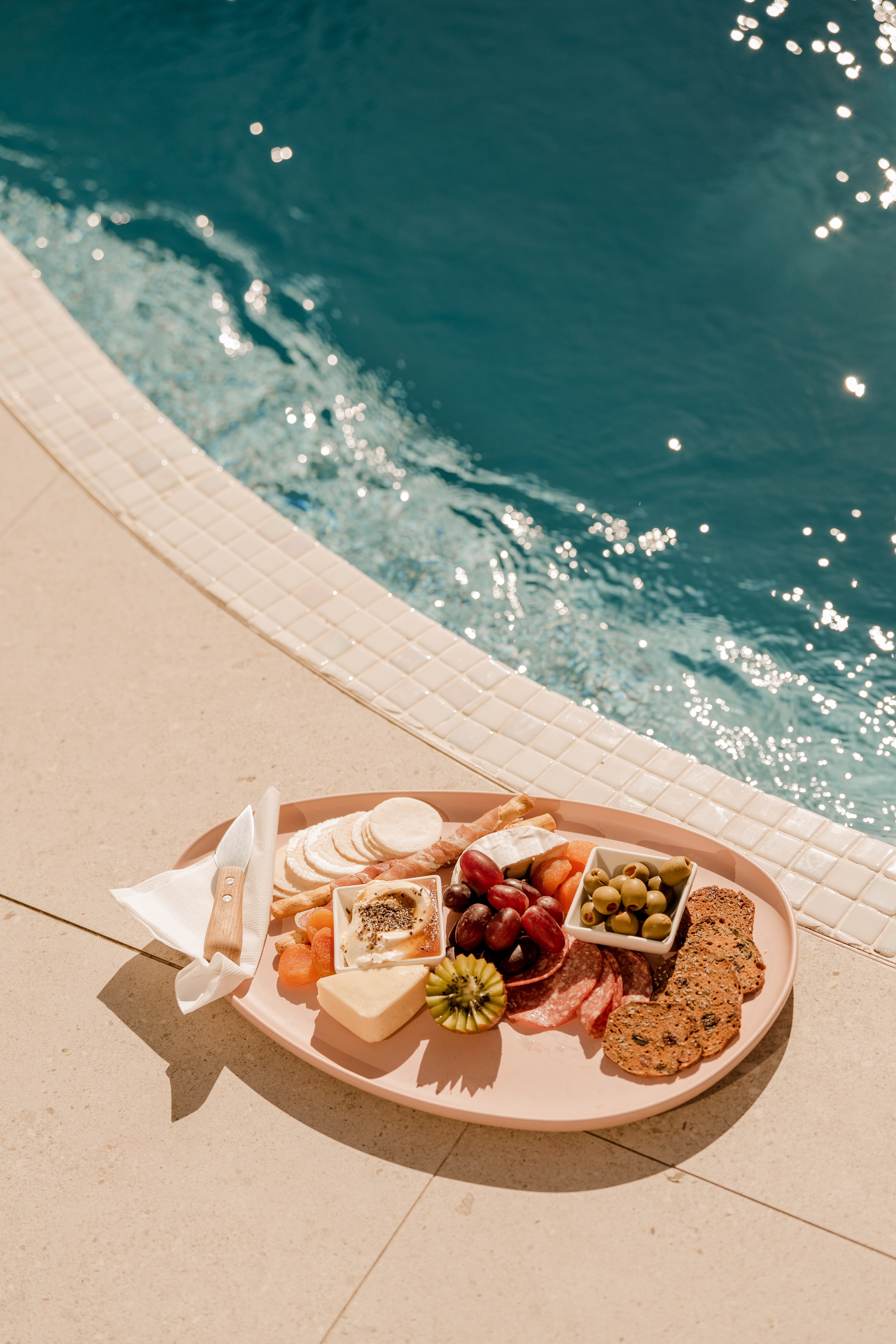 A pink grazing platter of cheeses, crackers, olives and charcuterie rests on the sunlit tiled edge of a sparkling pool.