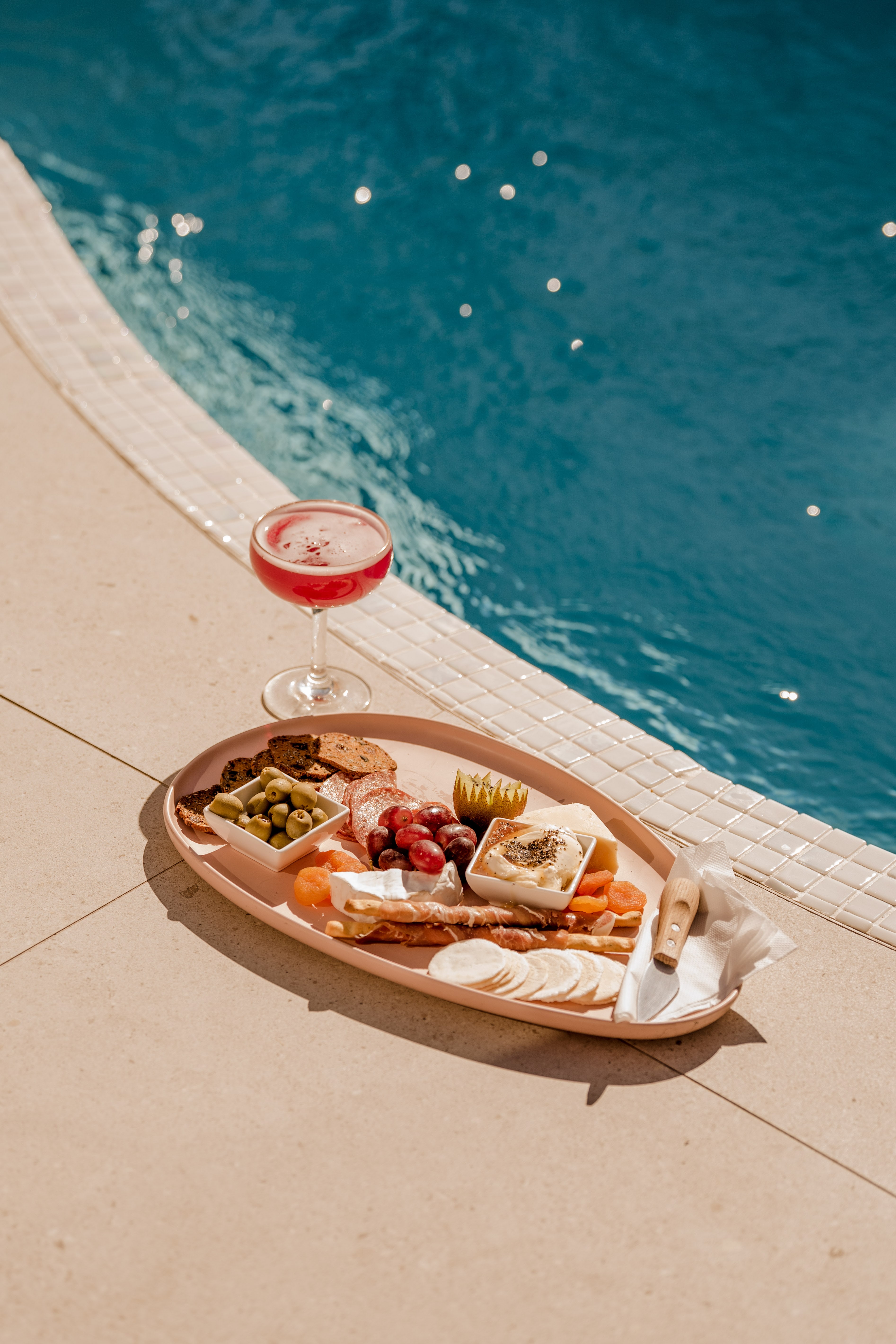 A pink oval grazing platter and a pink cocktail sit poolside on a sunny tiled deck beside shimmering blue water.