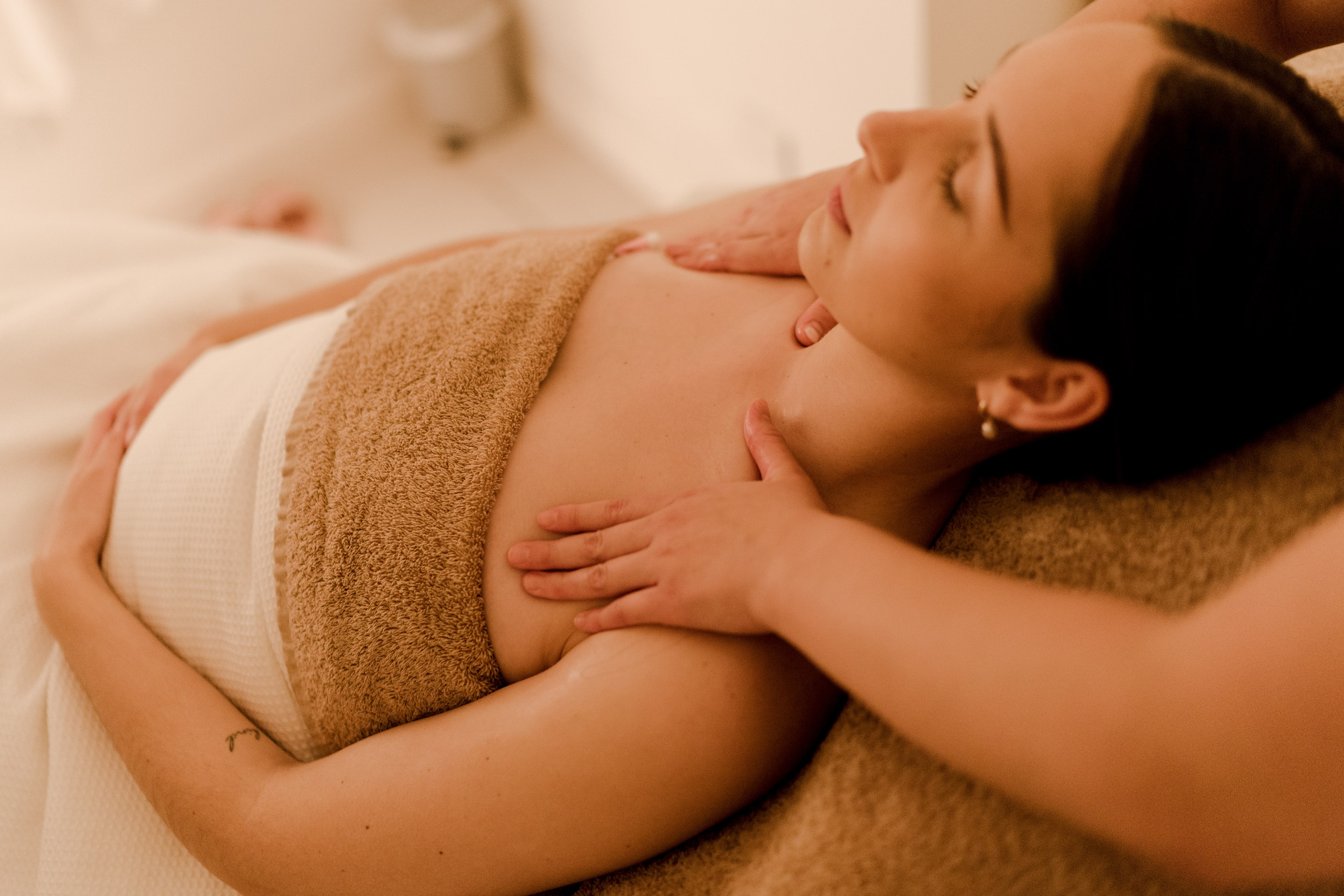 A woman lies wrapped in towels with eyes closed as a therapist massages her shoulders in a warmly lit treatment room.