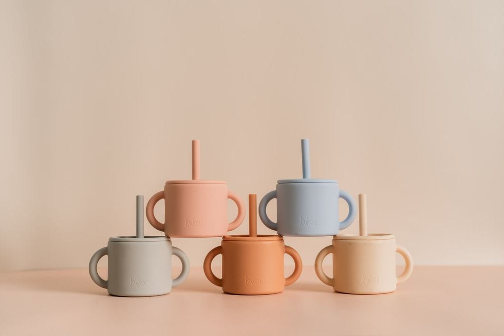 Five Bubsi silicone straw sippy cups with handles in grey, peach, blue, terracotta and cream, arranged in two rows on a soft pink surface.