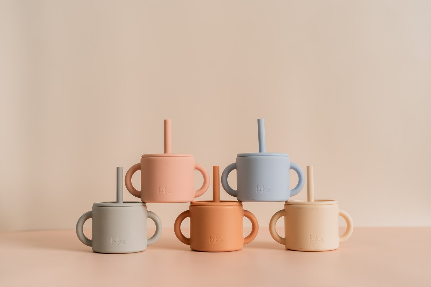 Five Bubsi silicone straw sippy cups with handles in grey, peach, blue, terracotta and cream, arranged in two rows on a soft pink surface.