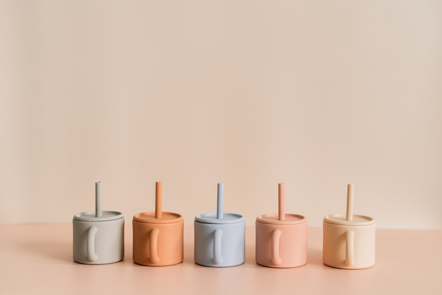 Row of five lidded Bubsi silicone straw cups with side handles in grey, terracotta, blue, peach and cream on a pink surface.