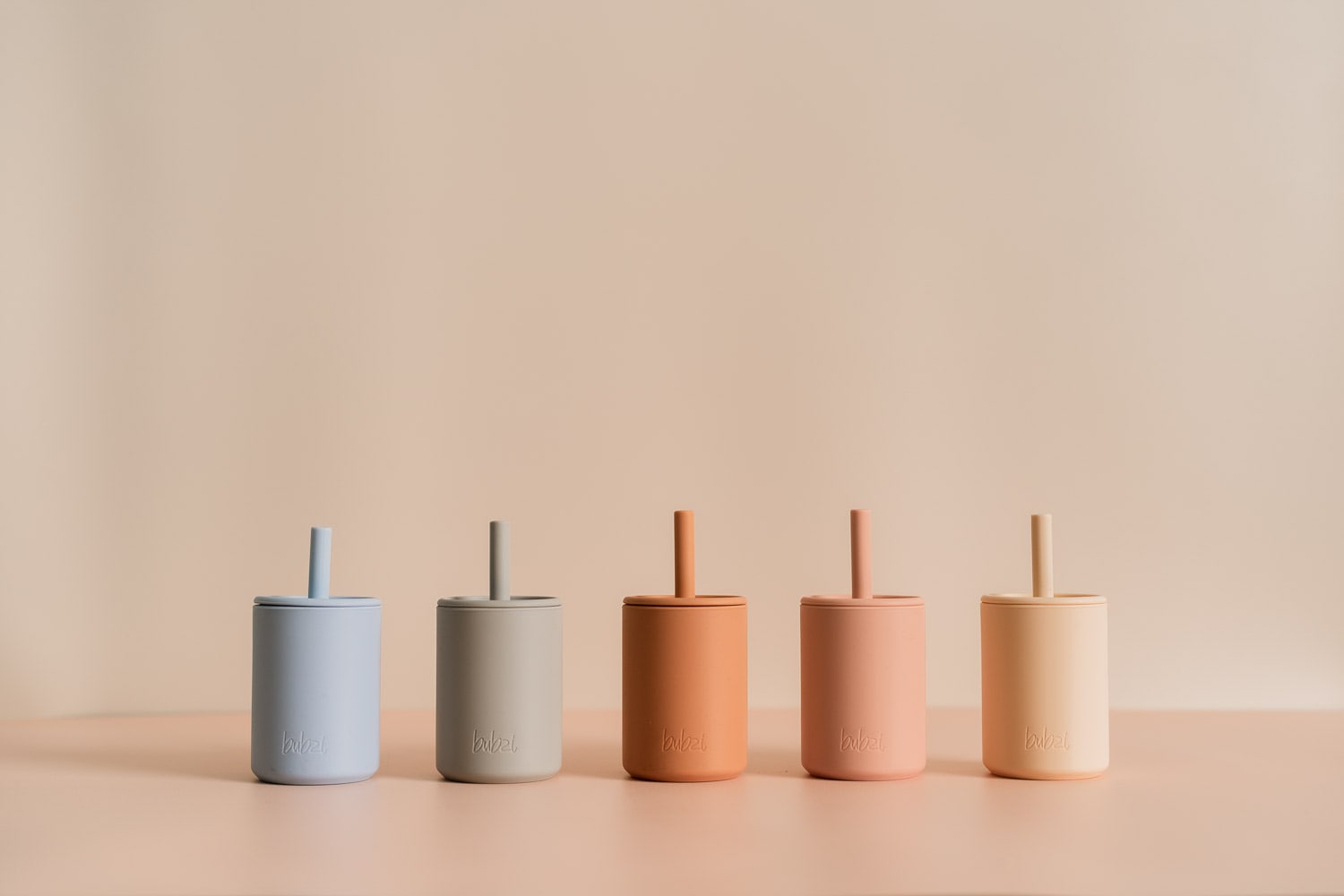 Five handleless Bubsi silicone straw cups with lids lined up in blue, grey, terracotta, peach and cream against a warm beige backdrop.