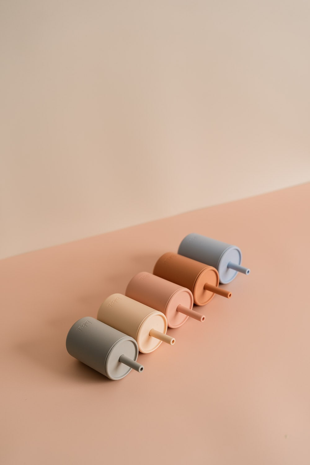Five Bubsi silicone straw cups laid on their sides in a diagonal row across a pink surface, in grey, cream, peach, terracotta and blue.