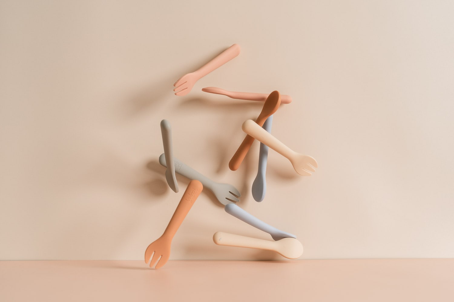 Bubsi children's silicone forks and spoons in peach, blue, grey, terracotta and cream scattered mid-fall against a soft beige background.