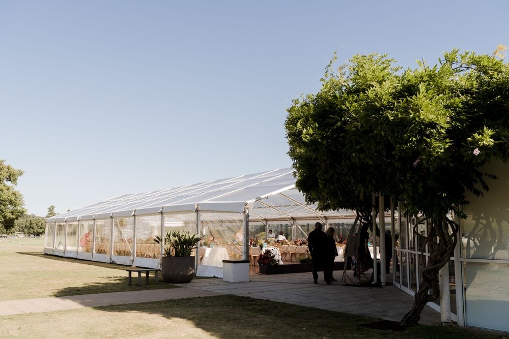 A large clear-roofed marquee set up for the Budget Petrol anniversary event on a sunny lawn, with dining tables inside and a leafy tree beside the entrance.
