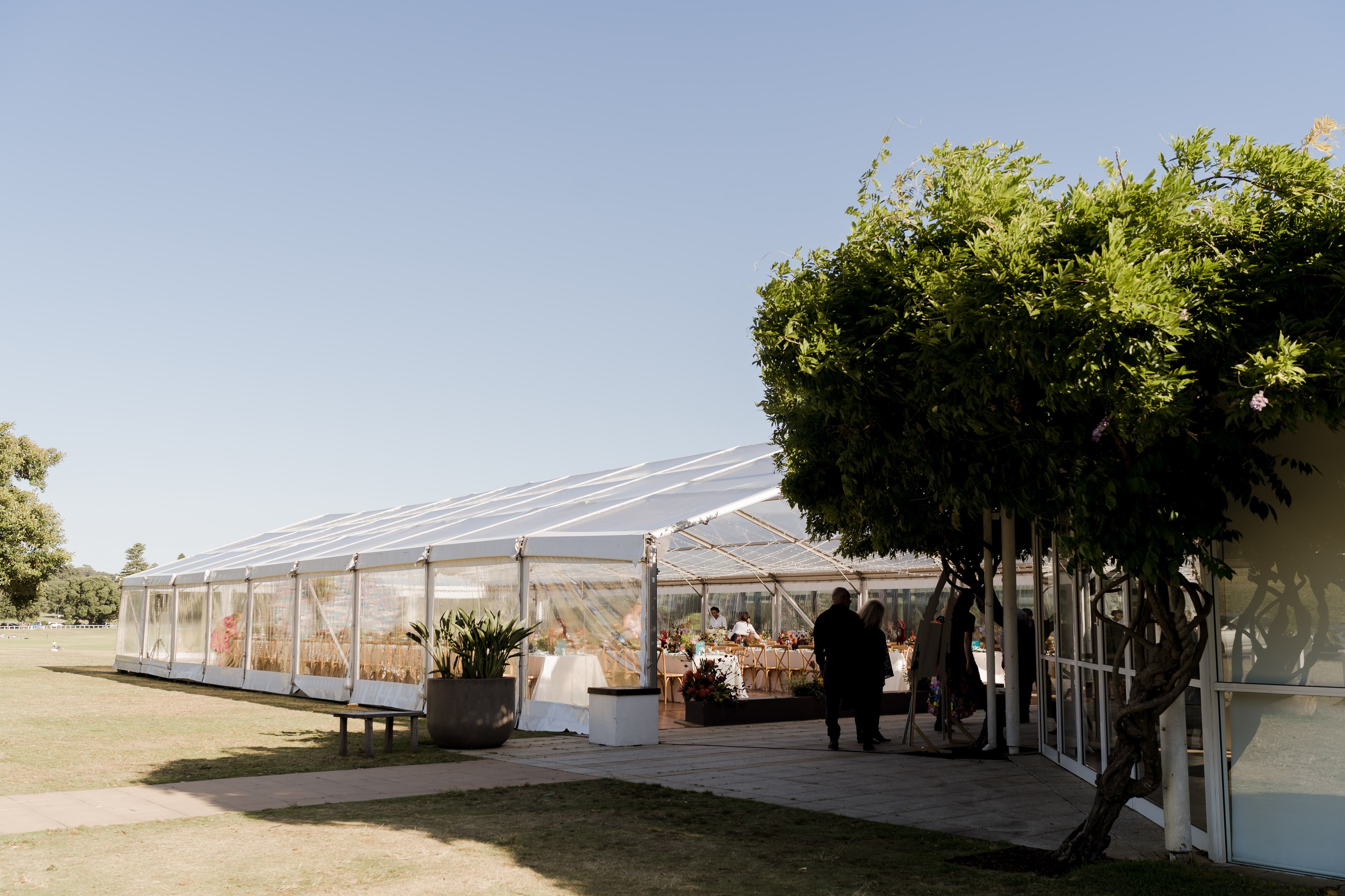 A large clear-roofed marquee set up for the Budget Petrol anniversary event on a sunny lawn, with dining tables inside and a leafy tree beside the entrance.