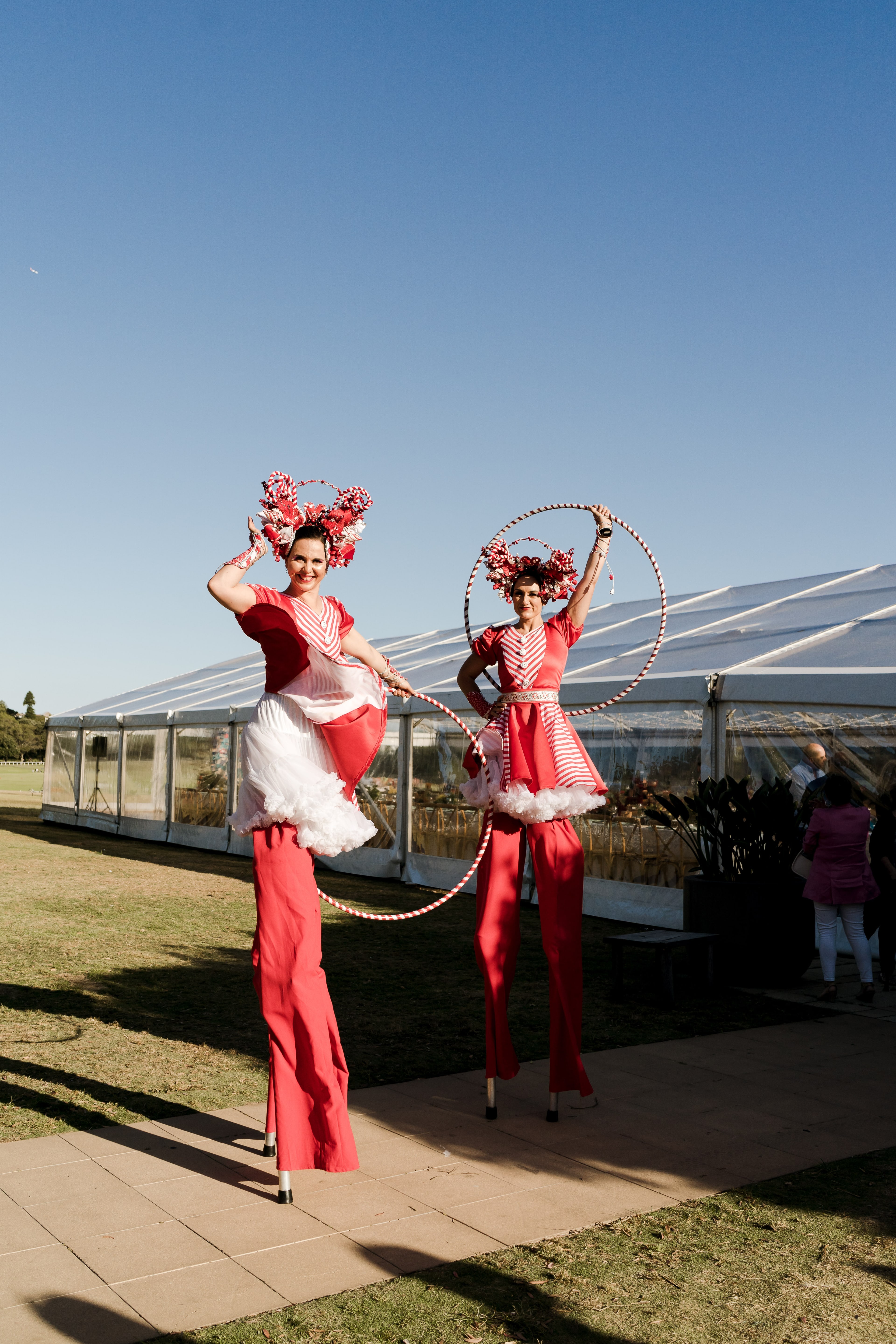 Two stilt-walking performers in red-and-white costumes twirl hoops outside the marquee under a clear blue sky at the anniversary celebration.