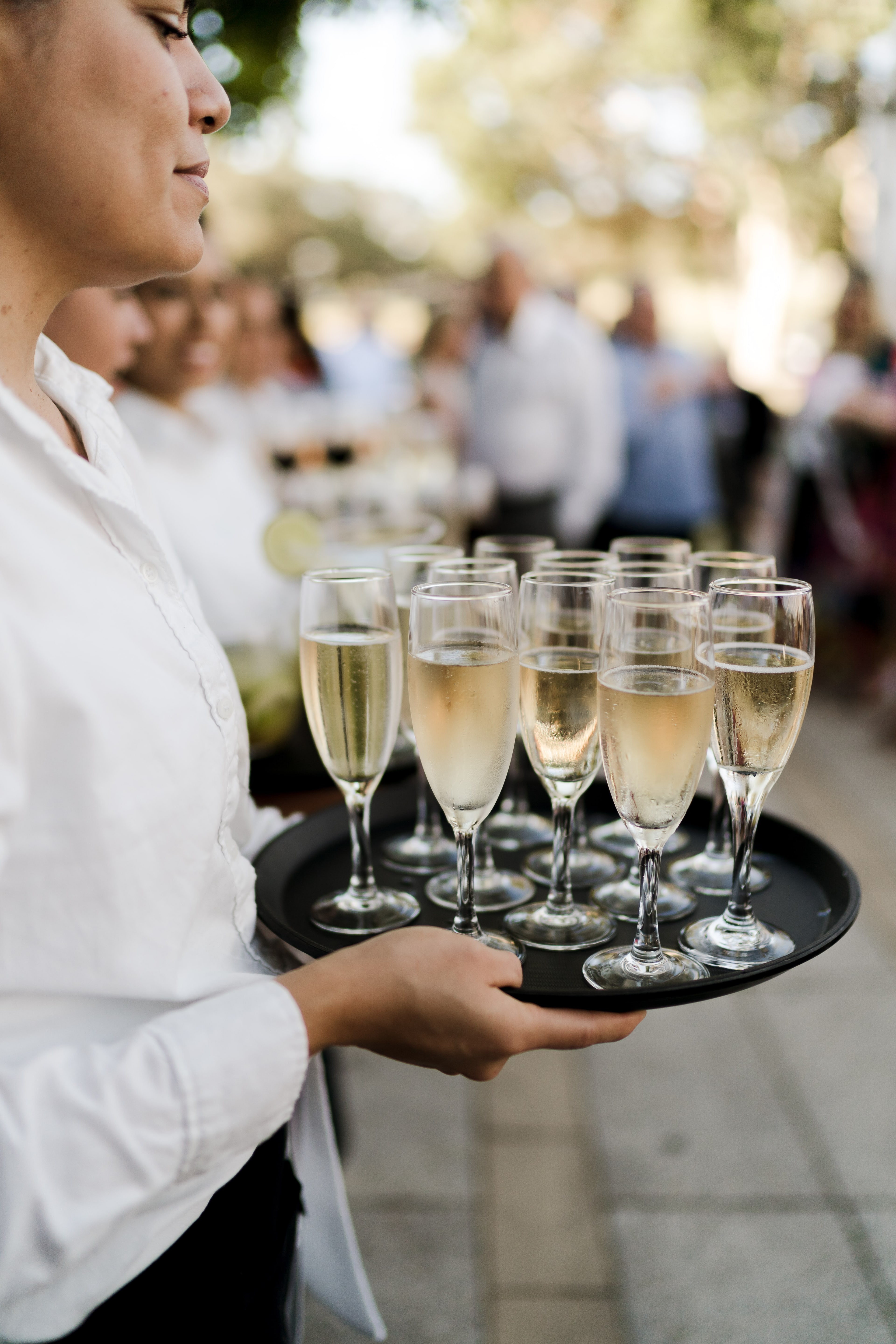 A waiter in a white shirt carries a black tray of filled champagne flutes to guests during outdoor drinks at the anniversary event.