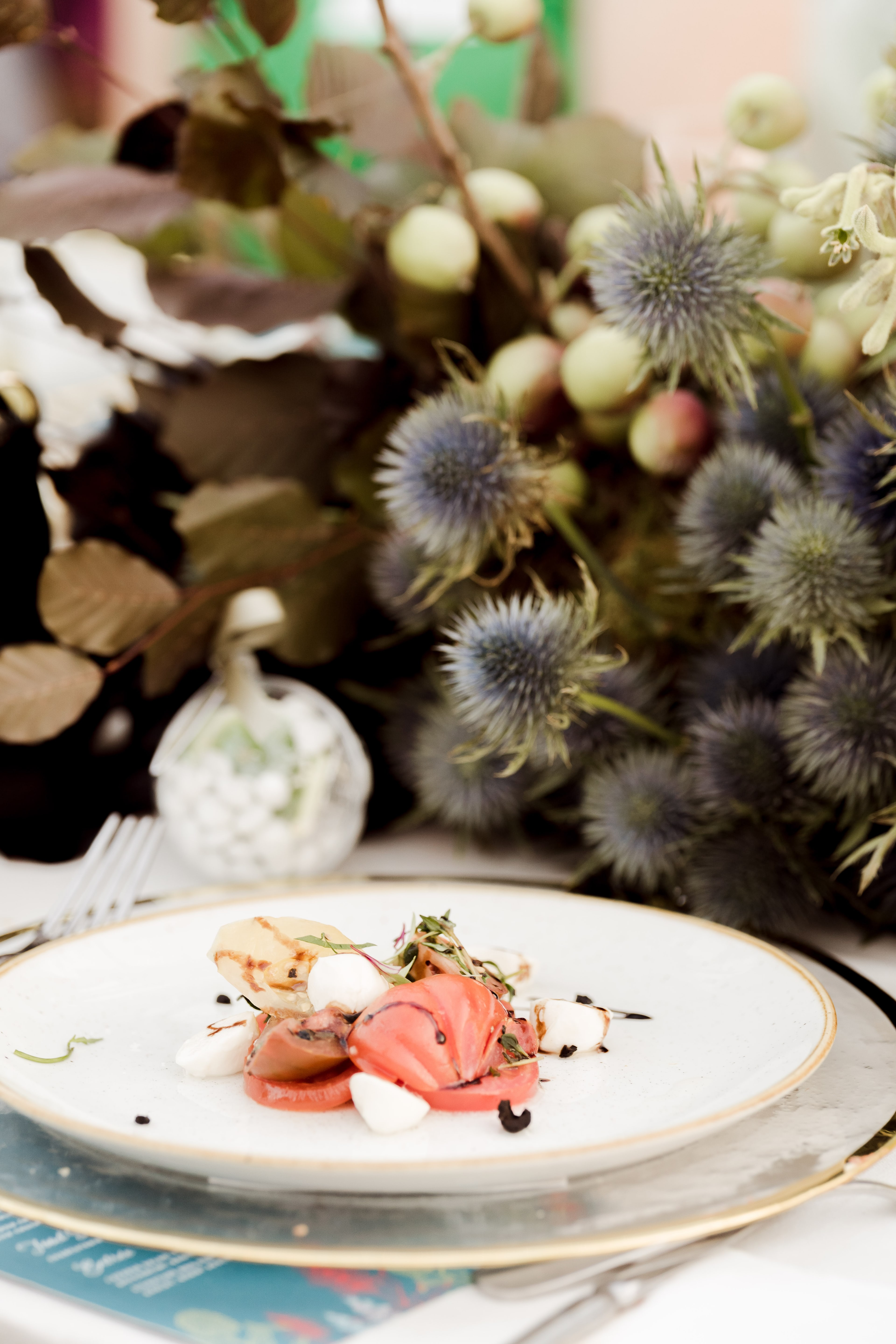 A plated tomato and bocconcini entree with balsamic on a gold-rimmed plate, styled beside a native floral arrangement with blue thistles.