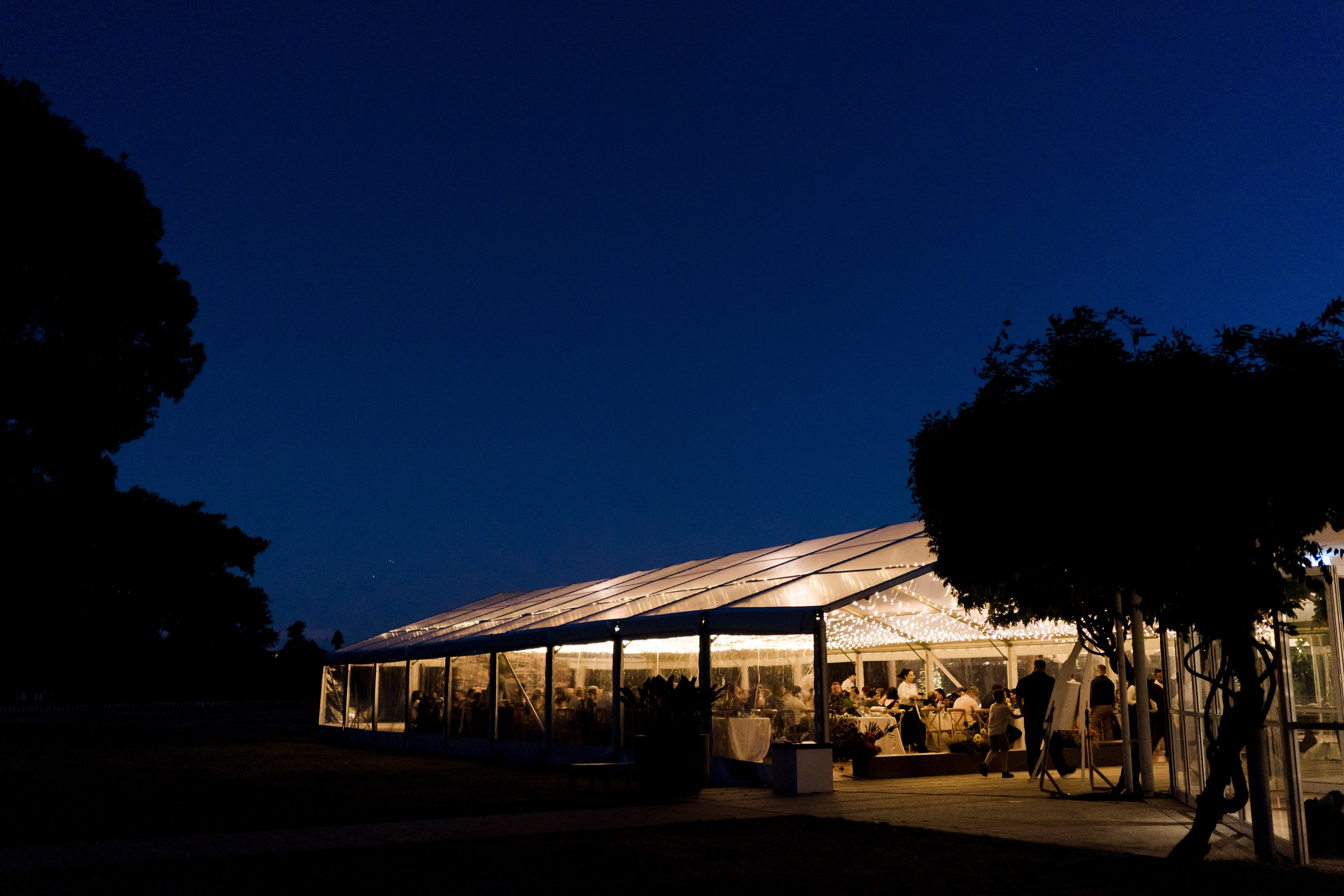 The clear marquee glows with warm light against a deep blue twilight sky as guests dine inside on the lawn at the anniversary event.