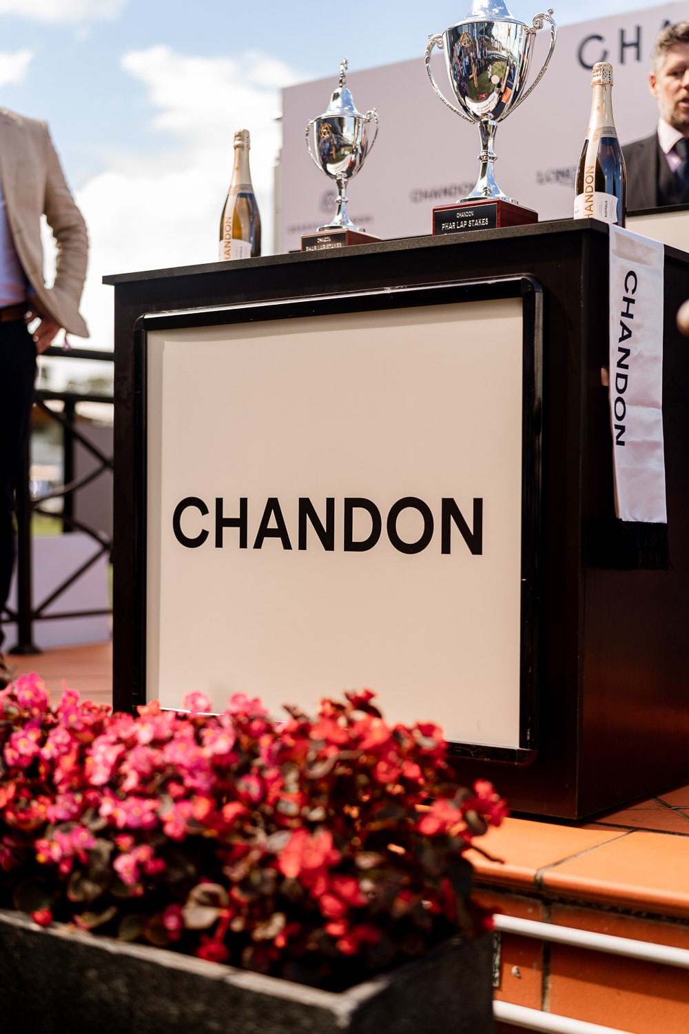 A branded Chandon plinth topped with silver Phar Lap Stakes trophies and sparkling wine bottles, with red flowers blooming in the foreground.