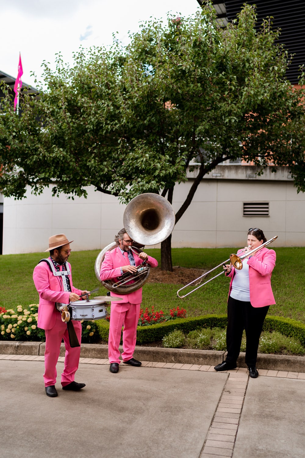 A three-piece brass band in matching pink suits plays sousaphone, trombone and snare drum on a garden path, entertaining guests outdoors.