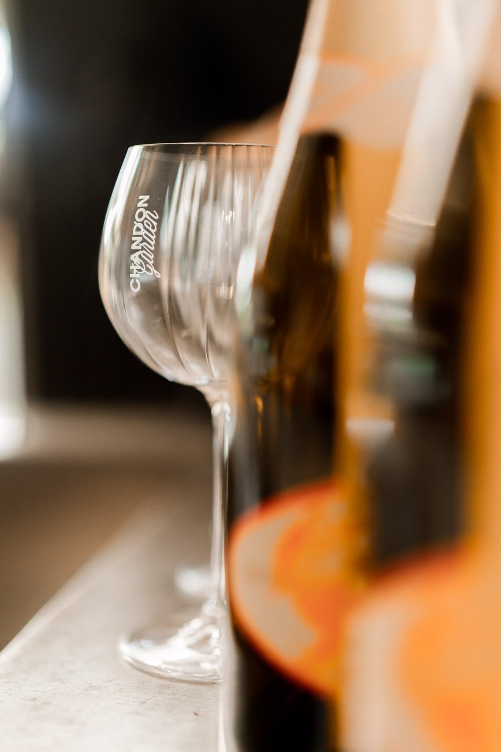 A close-up of an empty Chandon Garden Spritz glass on a stone bar top beside softly blurred bottles in warm sunlight.