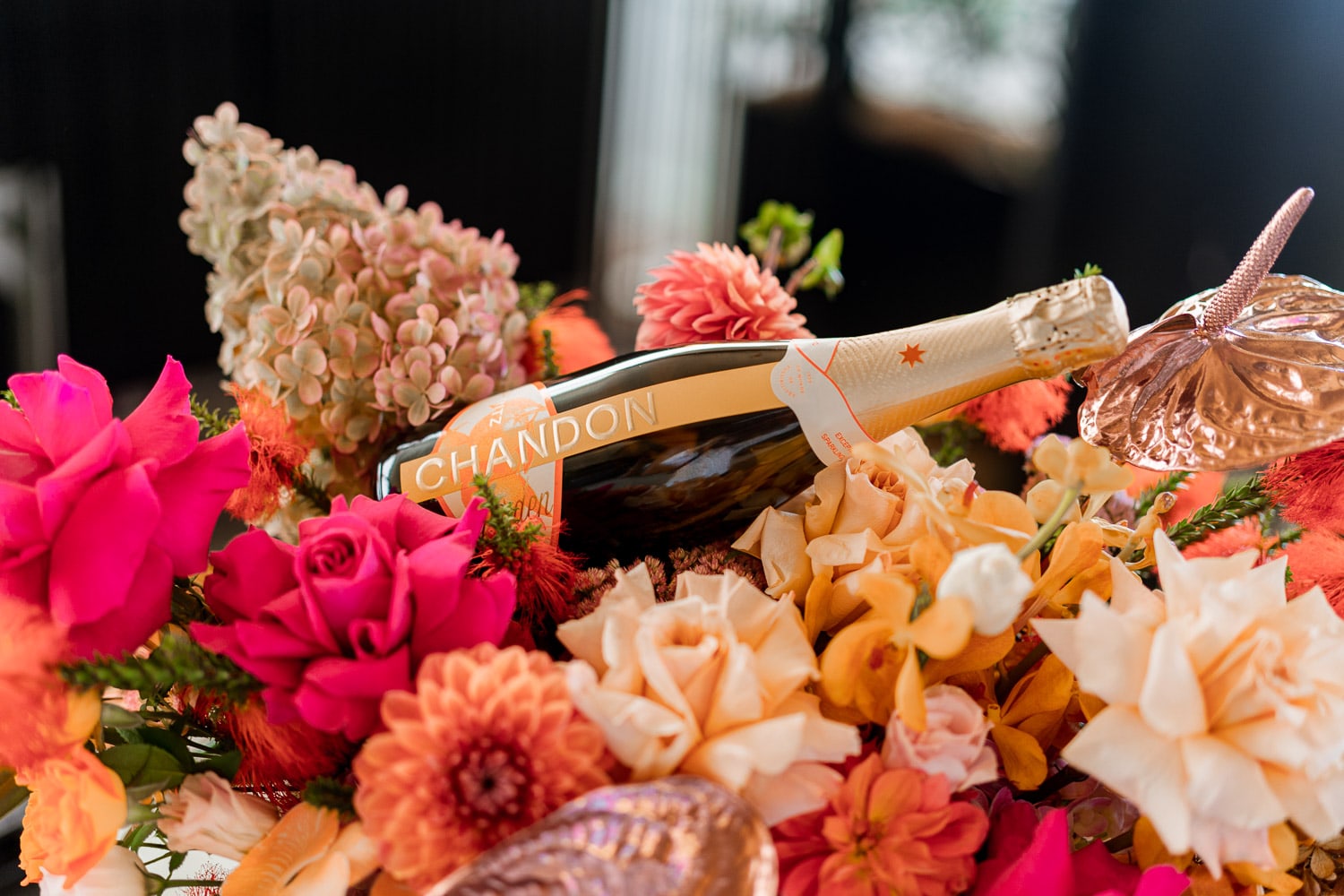 A bottle of Chandon sparkling wine nestled within a lush floral arrangement of pink roses, dahlias and orange blooms as a brand display.