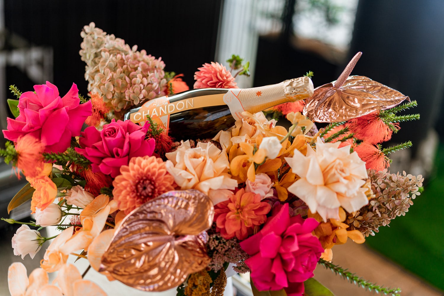 A Chandon sparkling wine bottle displayed amid an elaborate arrangement of pink and orange flowers with rose-gold anthurium accents.