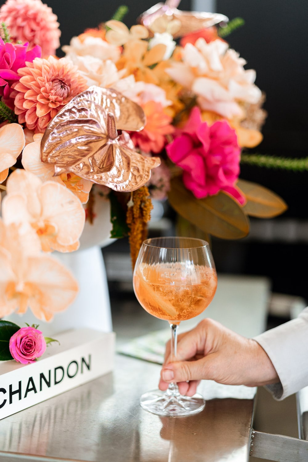 A guest's hand holds a Chandon Garden Spritz beside a vibrant floral centrepiece and a Chandon-branded book on a metal bar counter.