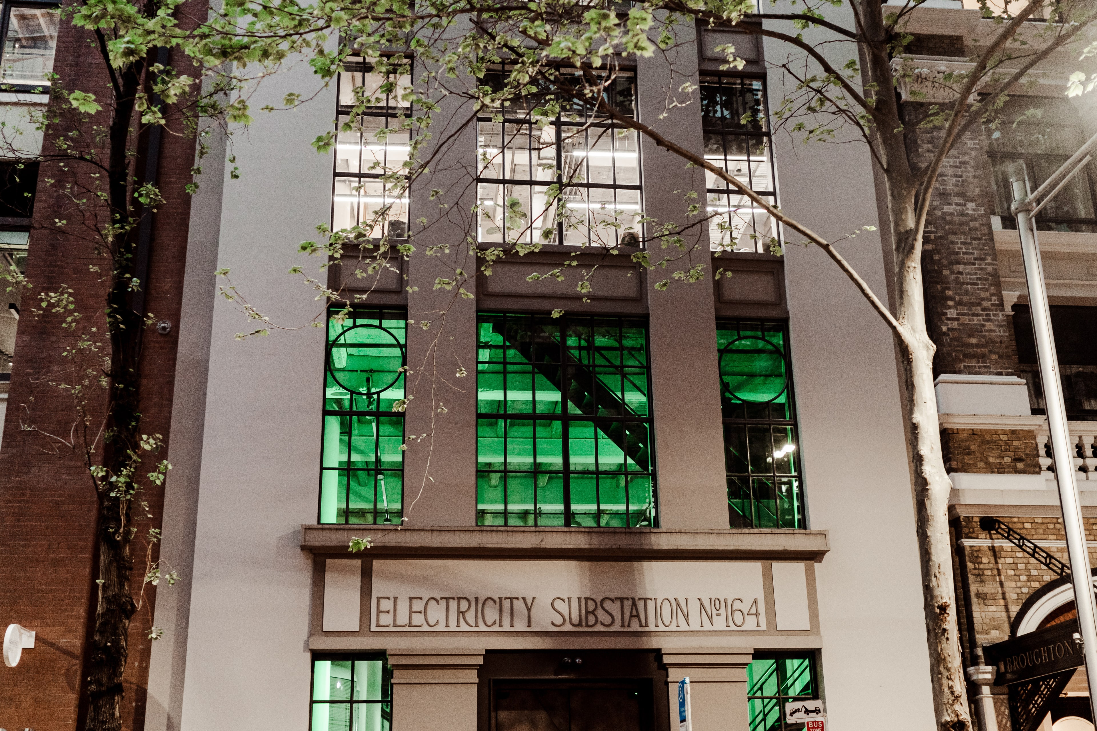 Heritage Electricity Substation No.164 building glowing green from within at dusk, the FC ClubHouse Sydney venue framed by bare street trees.