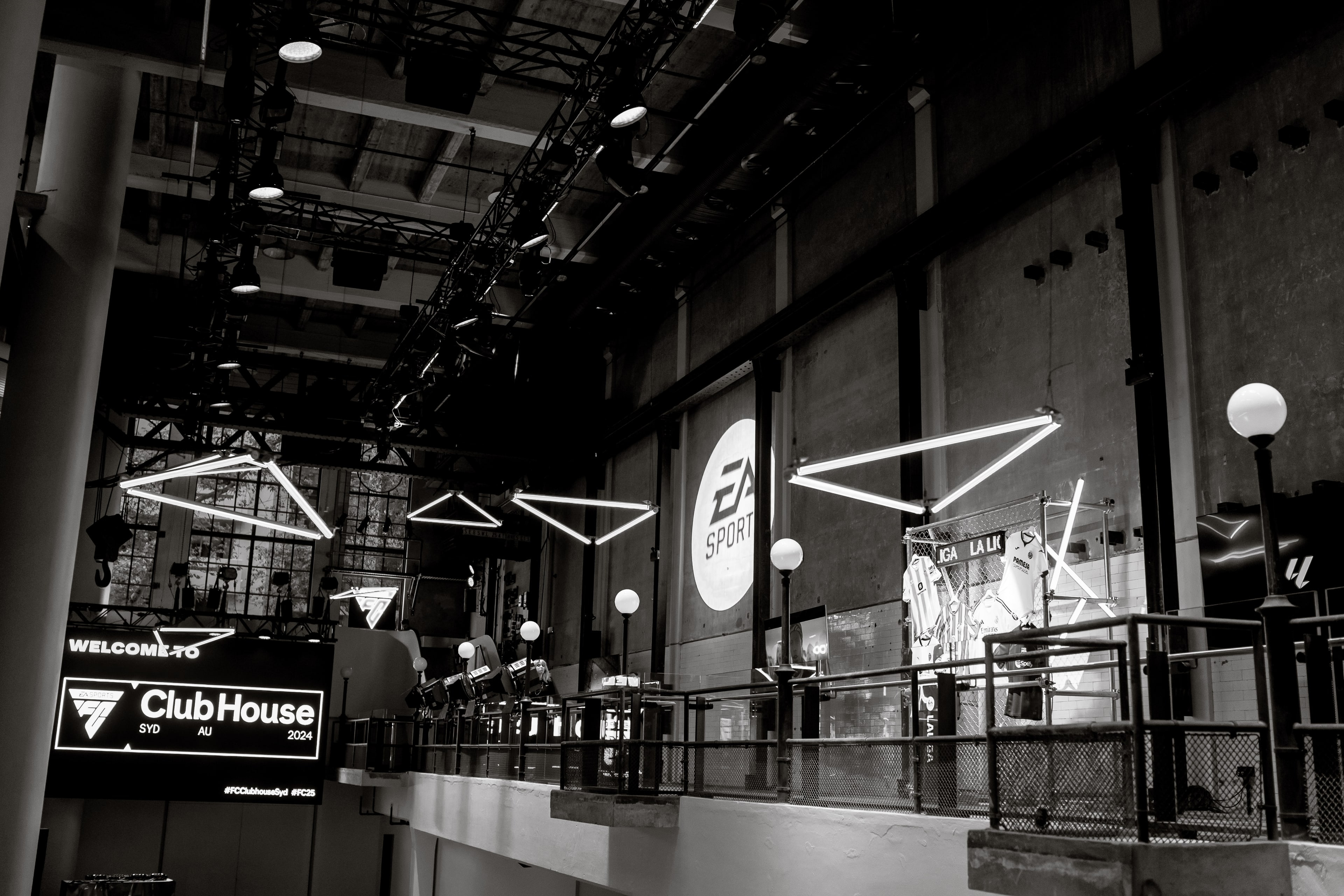 Black-and-white industrial venue interior with suspended geometric neon lights, an EA Sports logo wall and a lit 'Welcome to ClubHouse SYD AU 2024' sign.