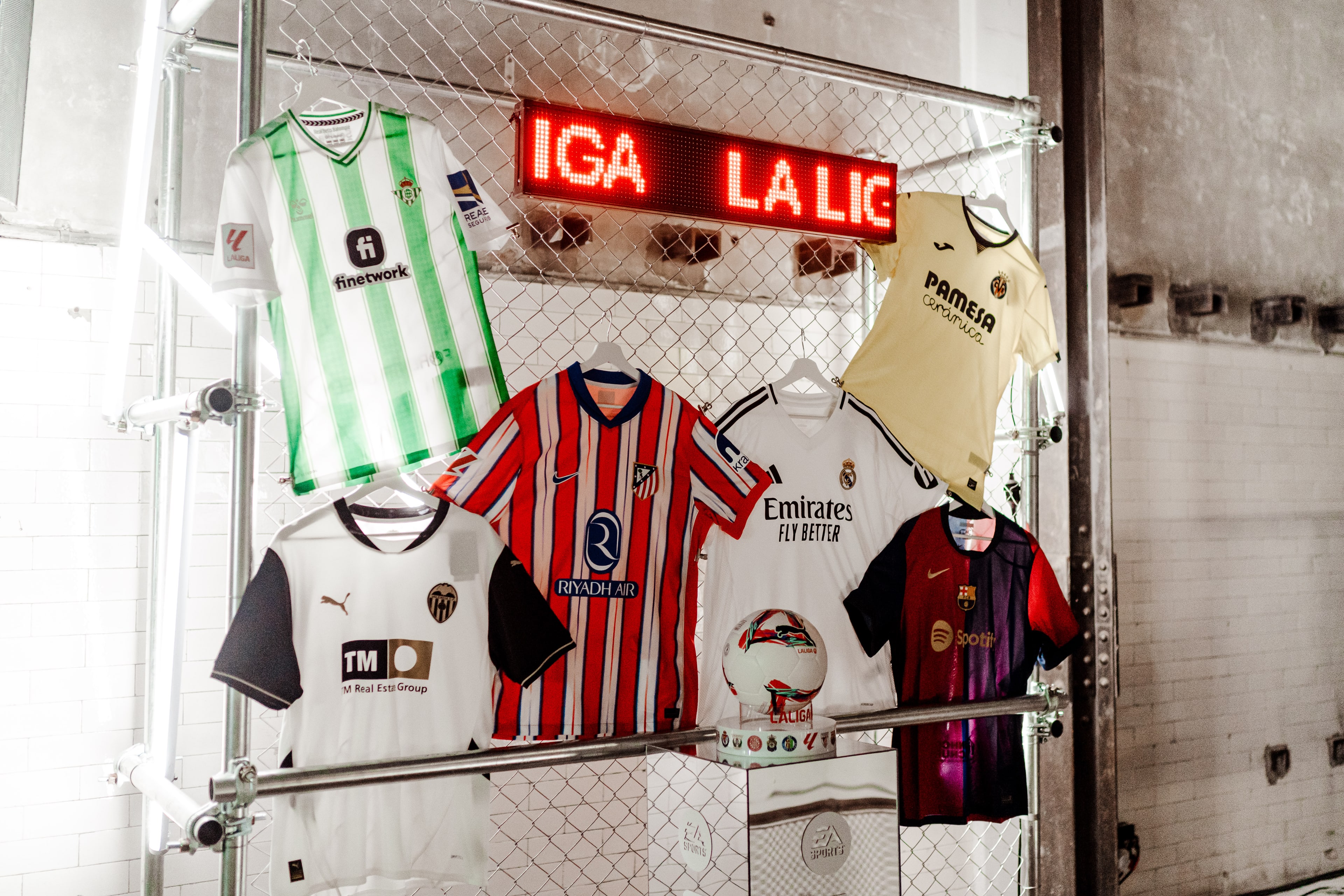 Football jerseys from Real Madrid, Barcelona, Atletico Madrid, Valencia, Betis and Villarreal hung on a wire fence beneath a red LED 'La Liga' sign.