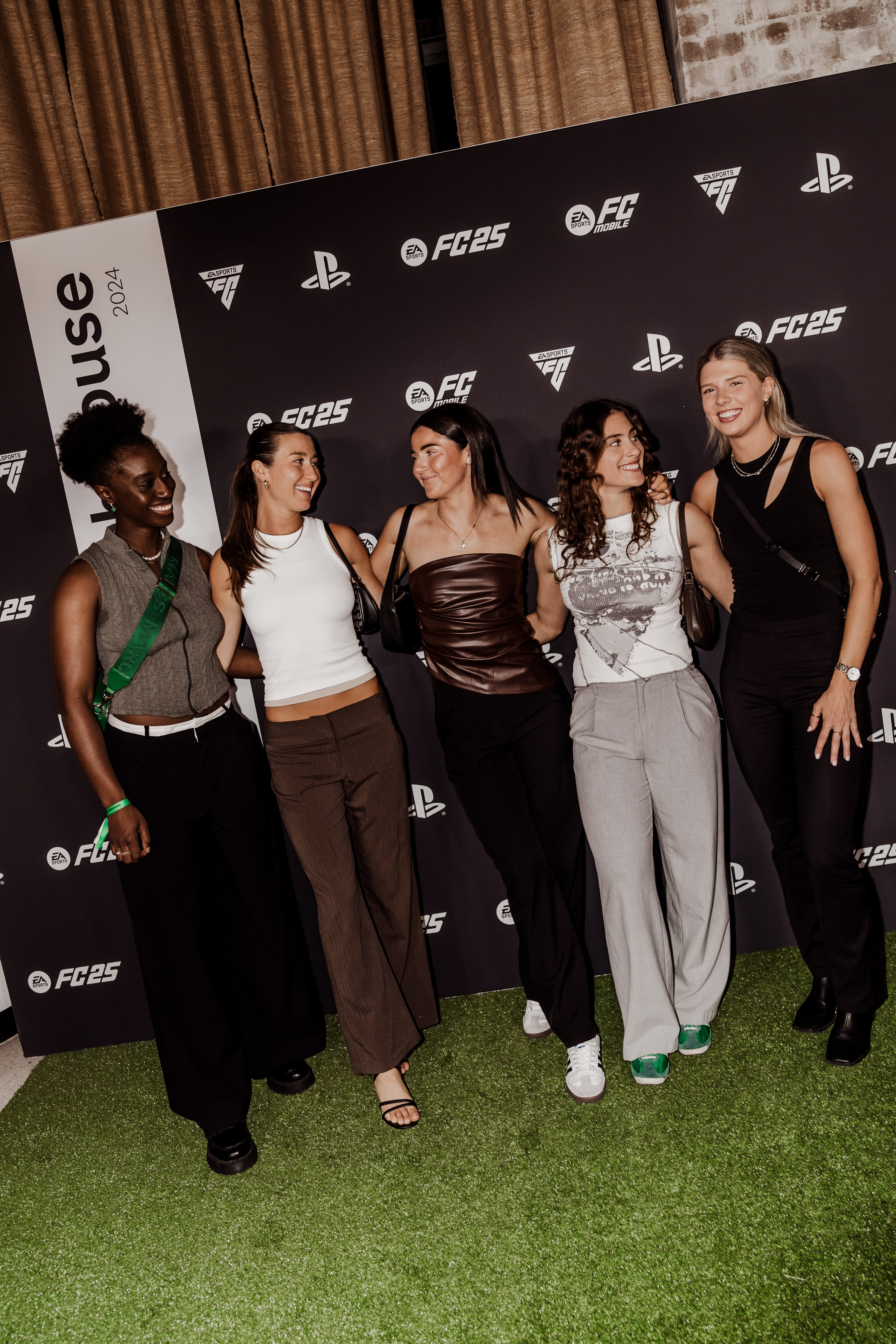 Five women pose and laugh together on green turf in front of an EA Sports FC 25 and PlayStation branded step-and-repeat wall.