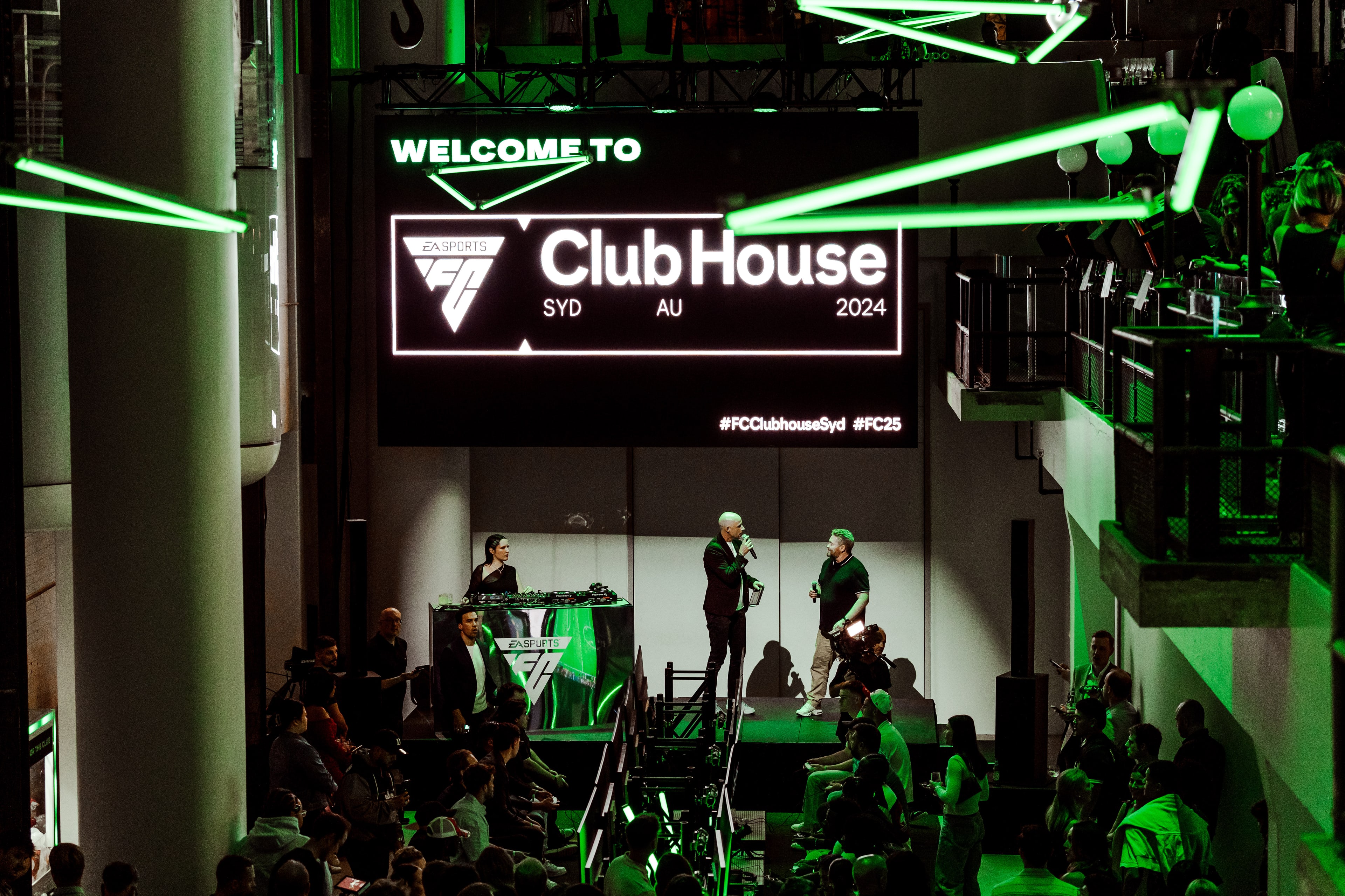 Crowd watches hosts on stage beneath a large 'Welcome to ClubHouse SYD AU 2024' screen amid green laser lighting in the darkened venue.