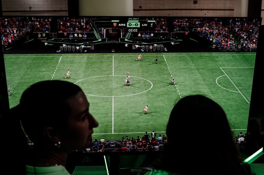 Two spectators in silhouette watch an EA Sports FC 25 Arsenal versus Liverpool match on a large screen, scoreboard reading 0-0 at 2:02.