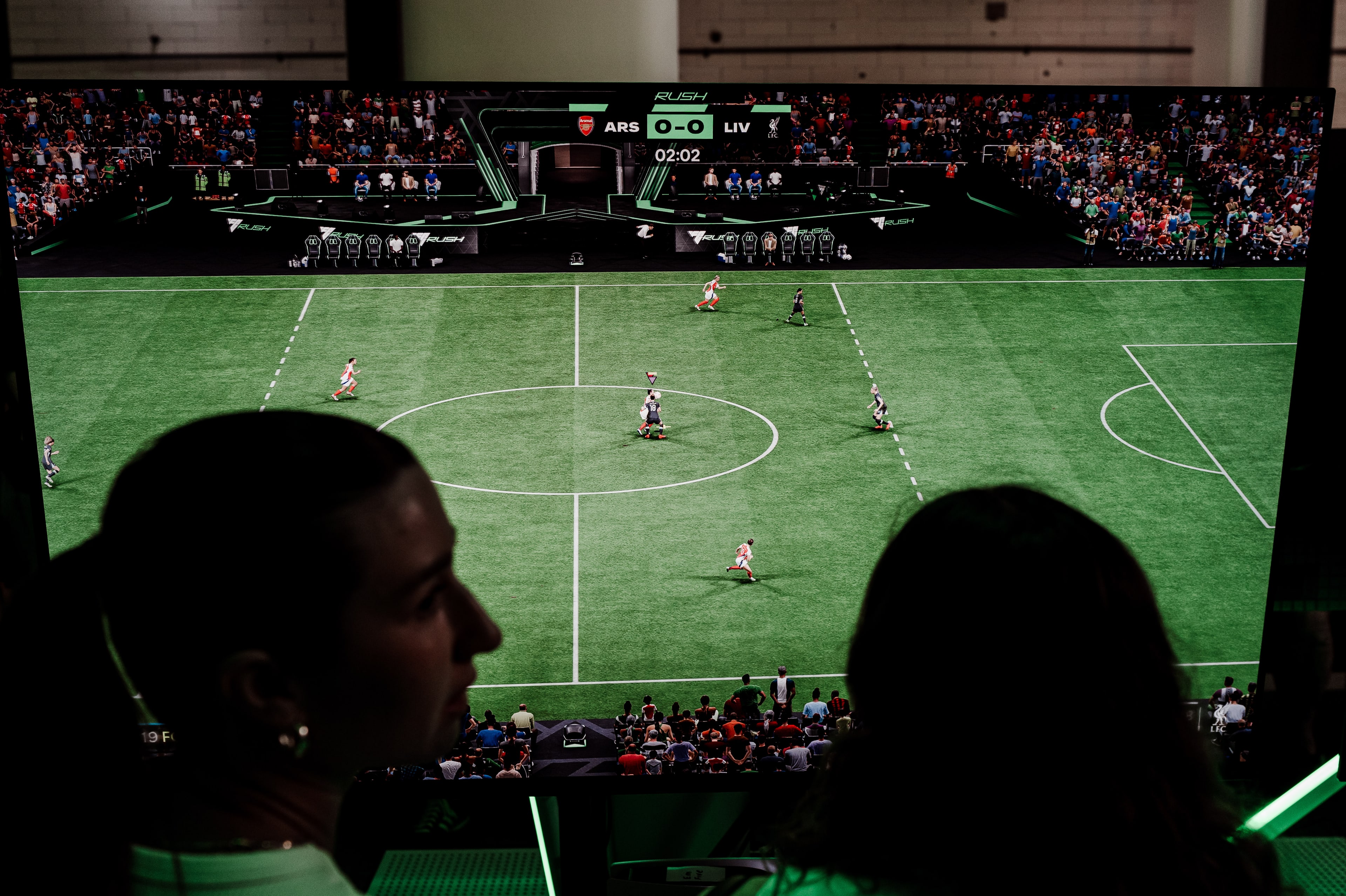 Two spectators in silhouette watch an EA Sports FC 25 Arsenal versus Liverpool match on a large screen, scoreboard reading 0-0 at 2:02.