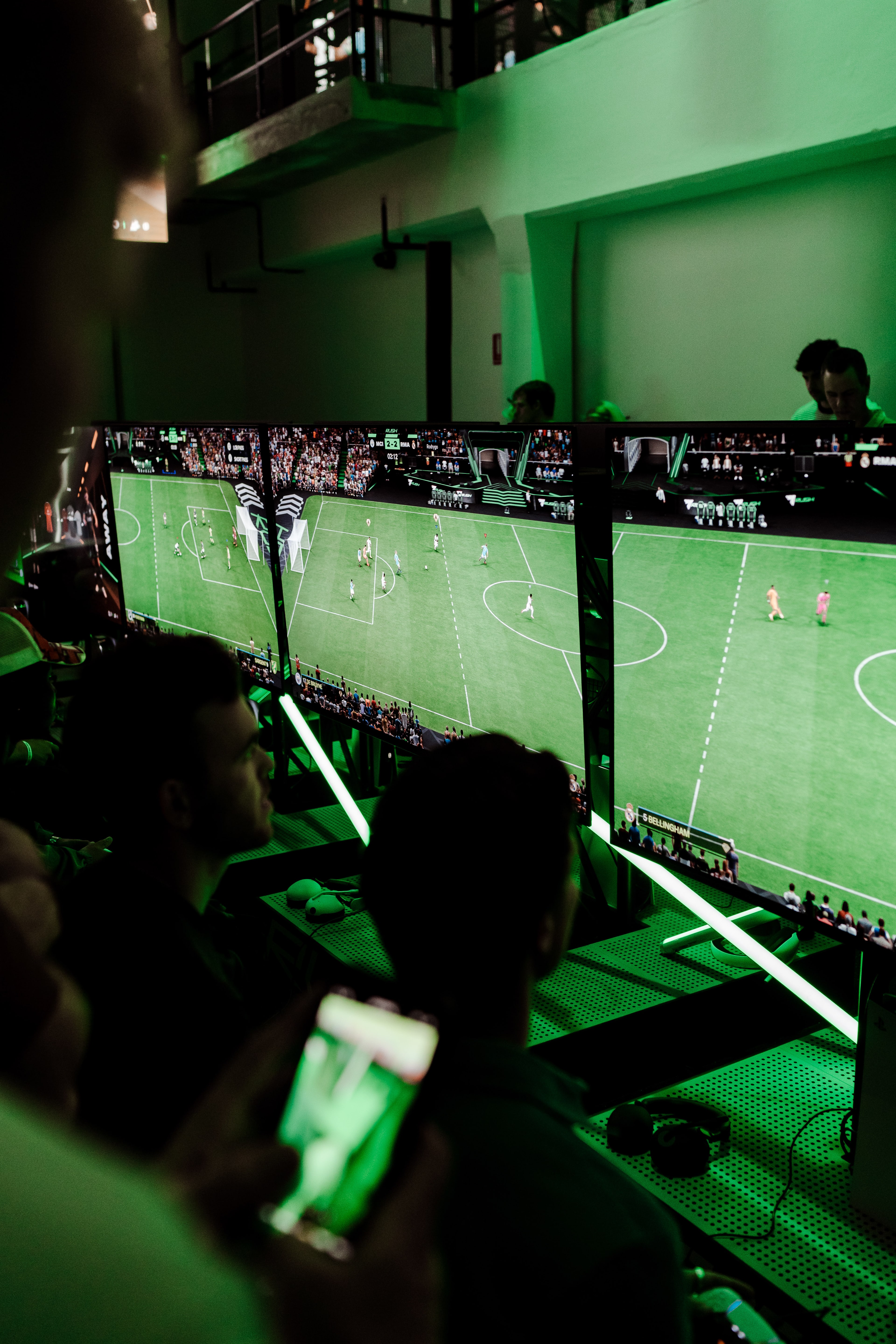 Attendees in green-lit gaming booths play EA Sports FC 25 across a row of monitors, one holding up a phone in the foreground.